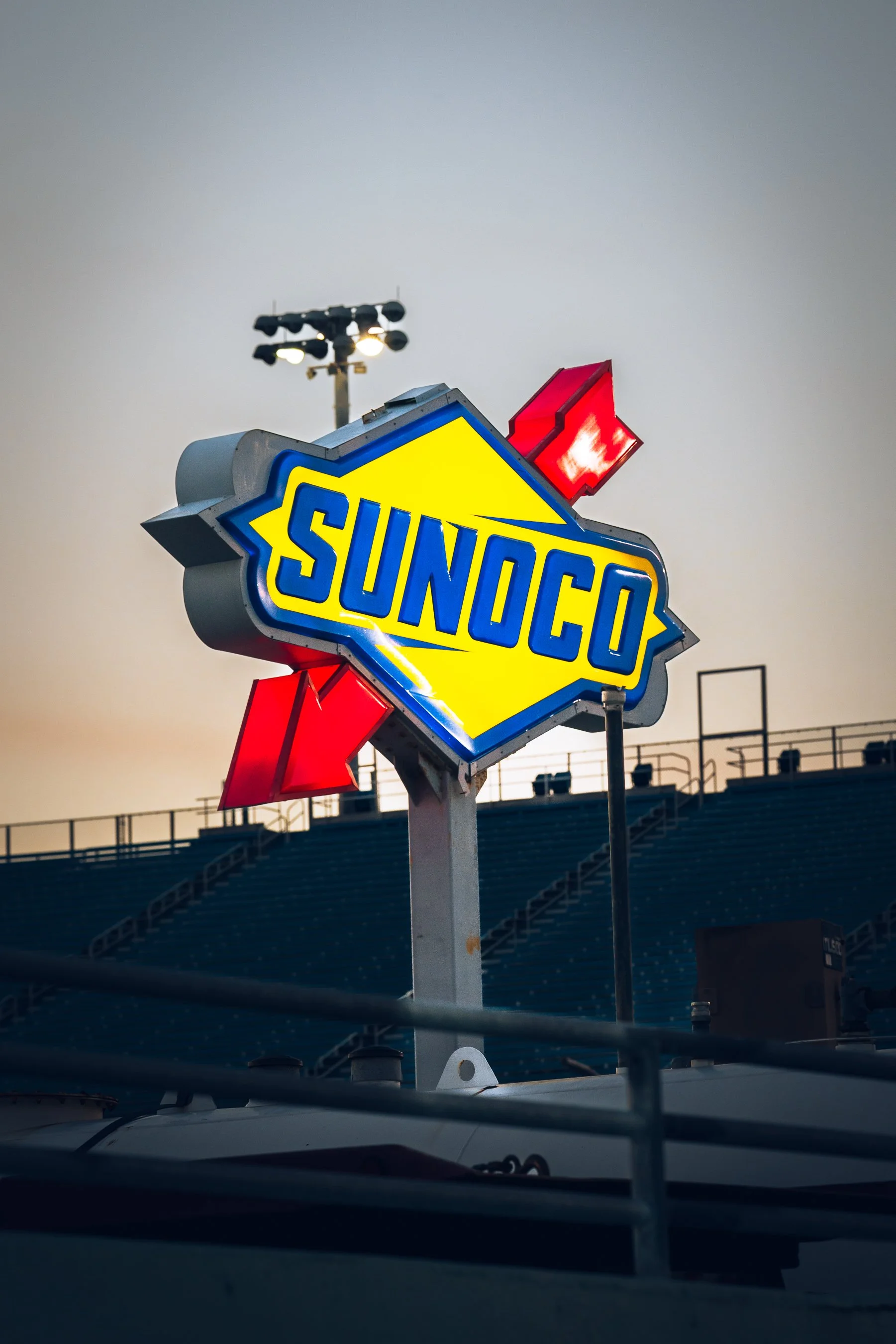 Sunoco gasoline station sign with yellow background and blue lettering, red directional arrows, and a light in the evening or early morning sky.