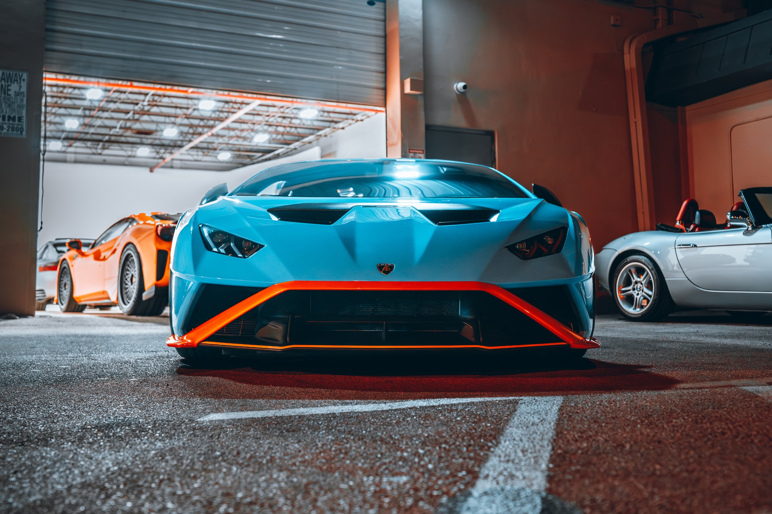 Front view of a blue Lamborghini sports car with orange accents parked in a garage with other luxury cars visible in the background.