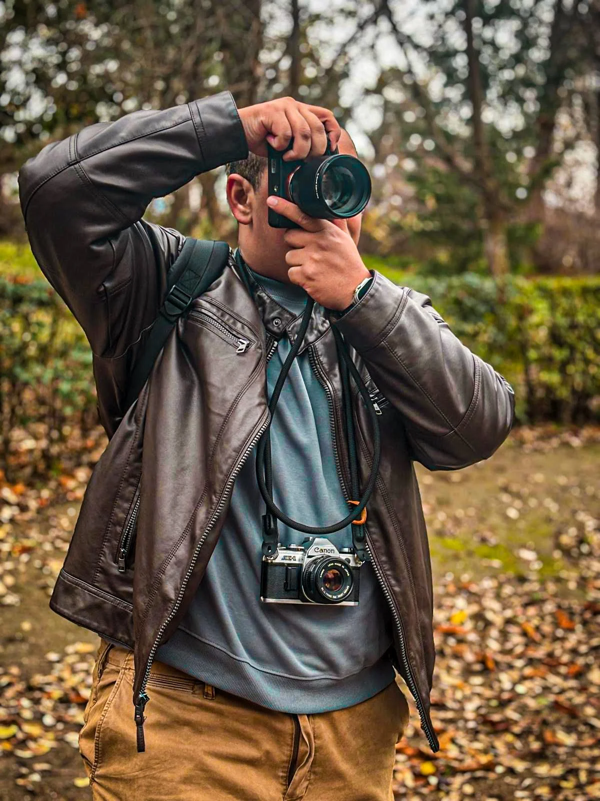 Person outdoors, wearing a brown leather jacket with a gray hoodie underneath, holding a camera up to their face, taking a photo. They have a camera hanging around their neck and another camera around their neck, standing on a path covered with fallen leaves, trees in the background.