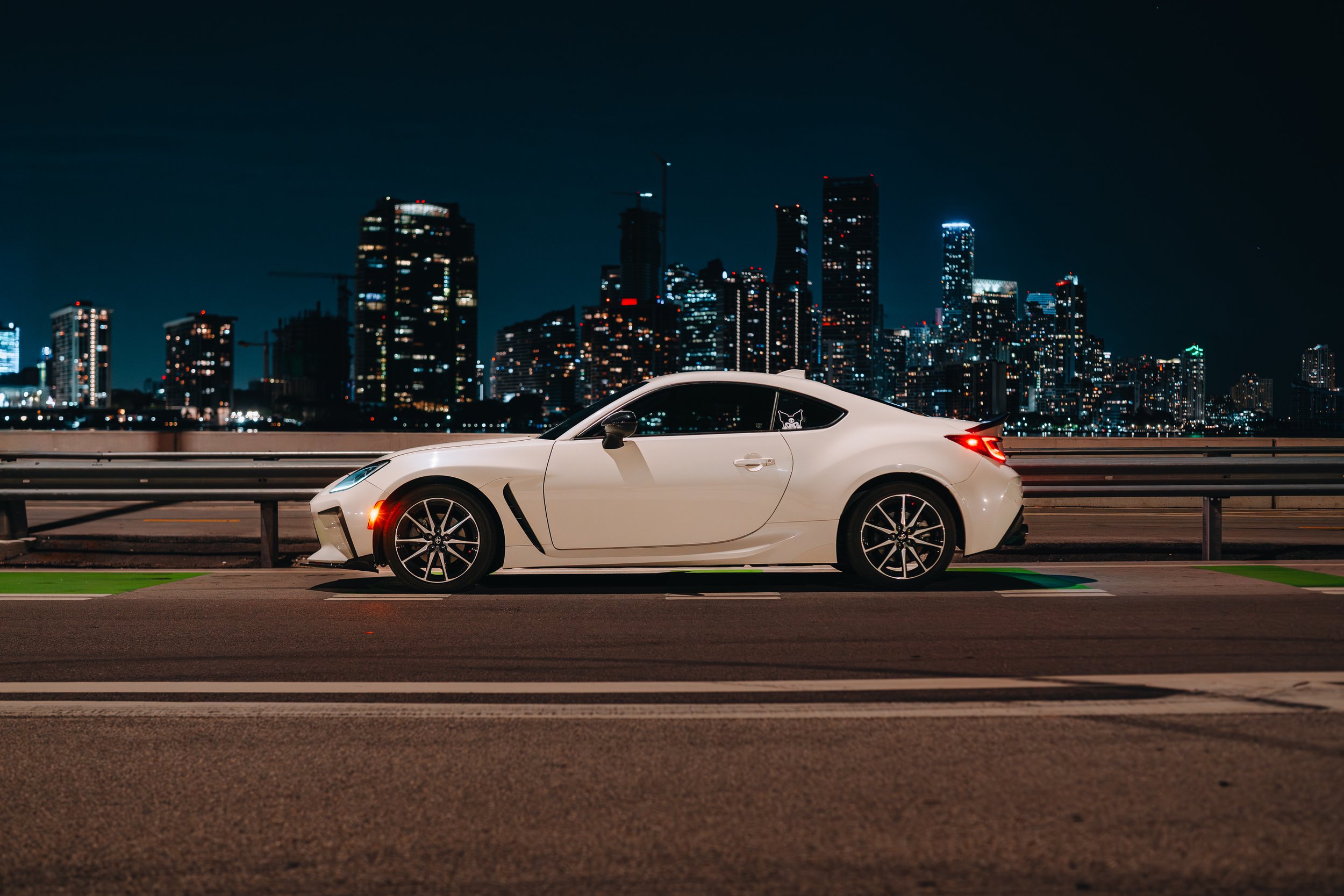 A white sports car parked on a road at night with a city skyline illuminated in the background.