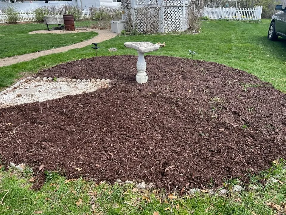 A garden bed with dark brown mulch, a white bird bath in the center, and small solar lights around it, surrounded by a well-maintained lawn and a picket fence in the background.