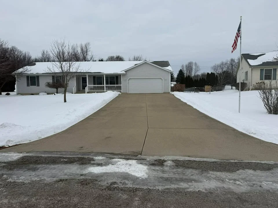 A snow-covered suburban house with a concrete driveway, a garage, an American flag, and leafless trees in winter.