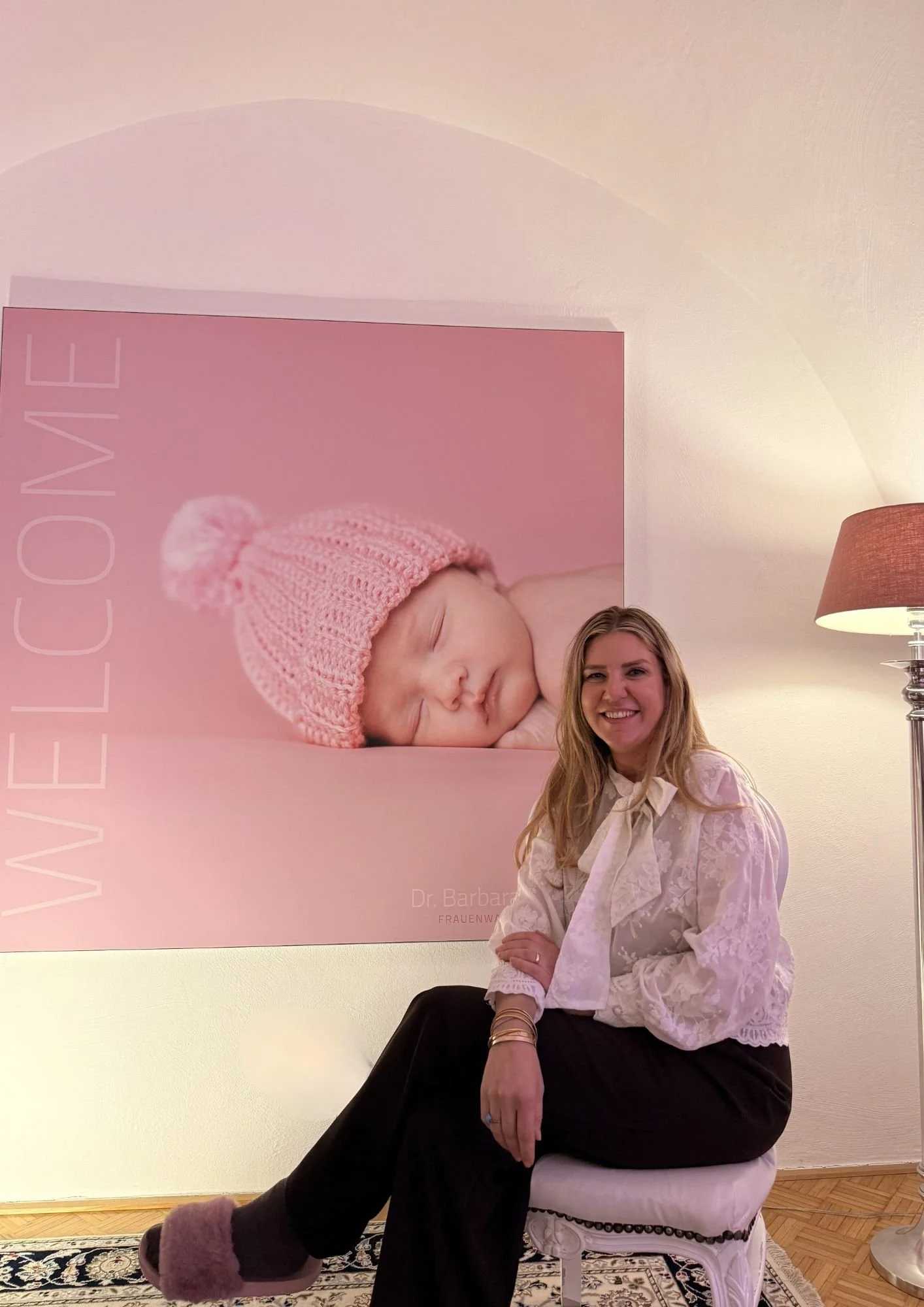 Eine Frau sitzt auf einem weißen Stuhl vor einem Wandbild eines schlafenden Babys mit rosa Mütze. Das Wandbild enthält den Text "LOVE" und den Namen "Dr. Barbara".