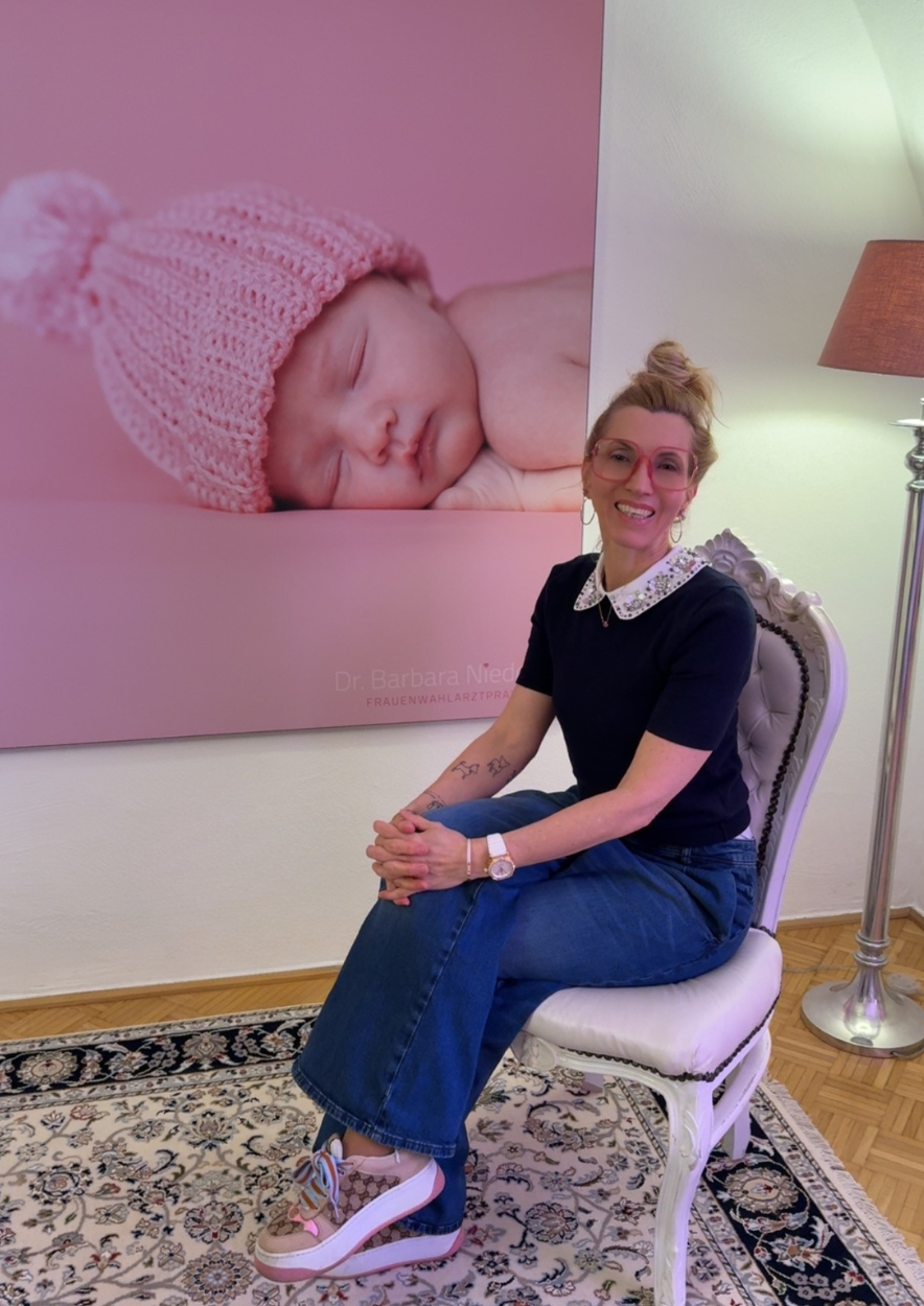 Frau sitzt auf einem weißen Stuhl vor einem Gemälde eines schlafenden Babys mit rosa Mütze. Sie trägt eine schwarze Bluse mit weißen kragen, eine Jeans und Turnschuhe. Im Hintergrund steht ein Regal und ein Lampenschirm.