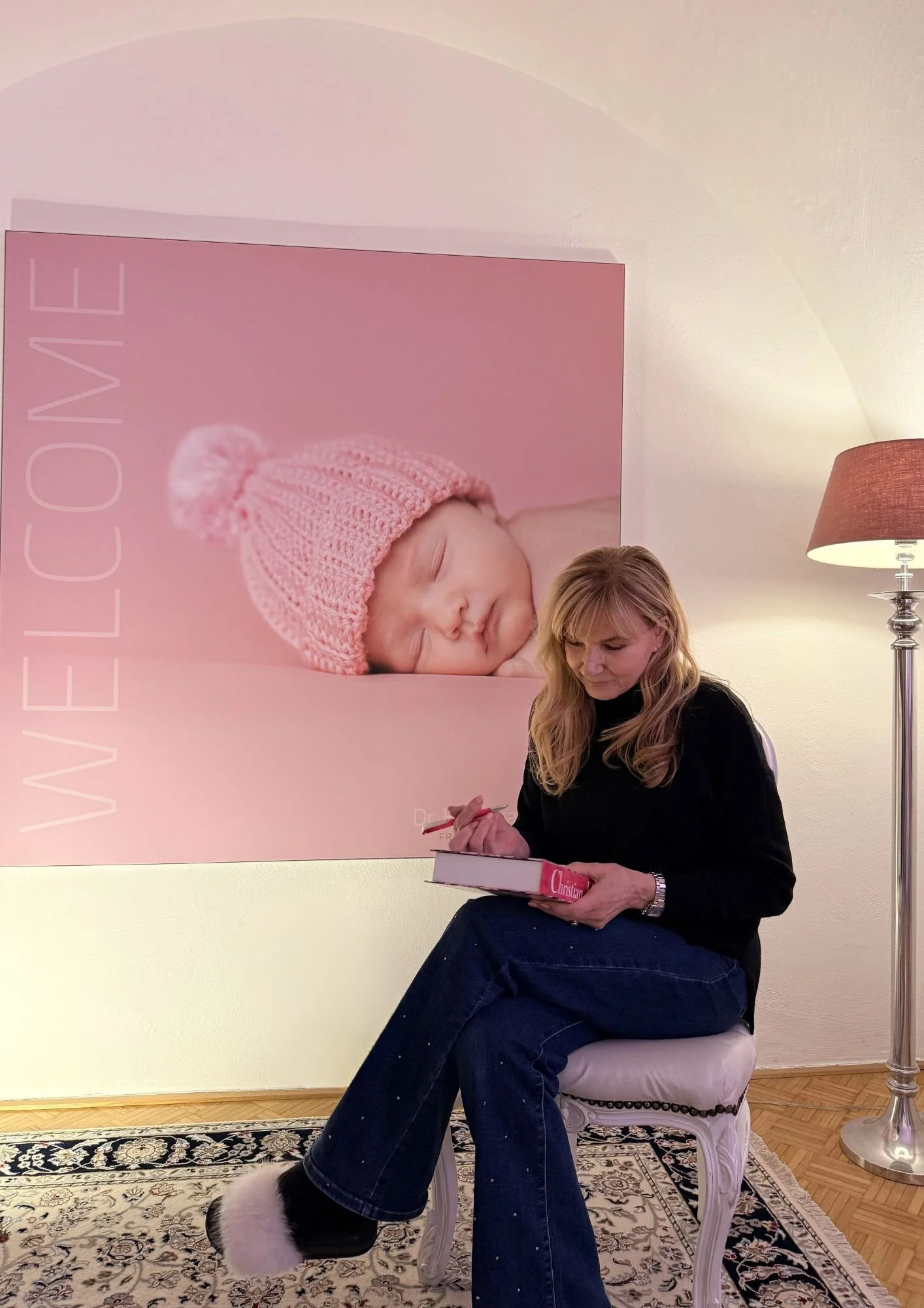 Eine Frau sitzt auf einem weißen Stuhl in einem Raum mit einem hellen Wand und steht vor einem großen Bild eines schlafenden Babys mit rosa Mütze. Die Frau liest in einem pinkfarbenen Buch.