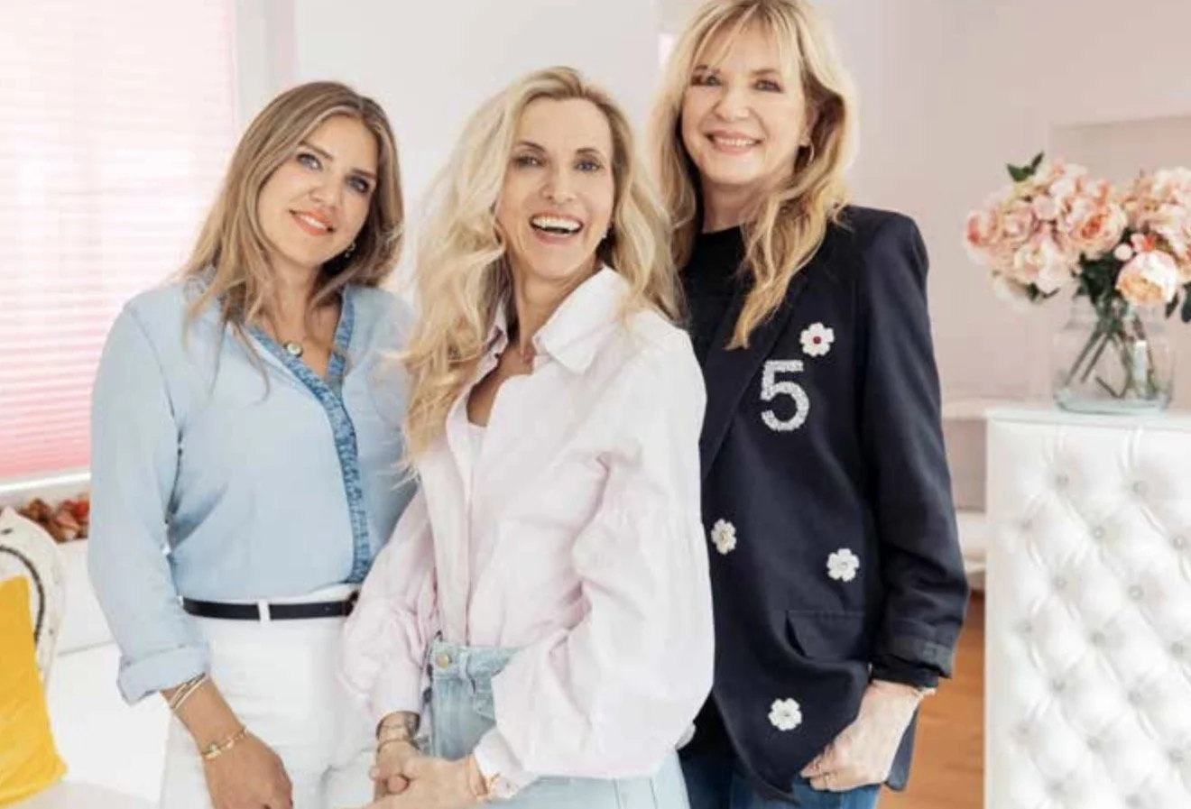 Drei lachende Frauen stehen nebeneinander in einem hellen Raum mit Blumen. Sie tragen modische Kleidung, eine trägt eine blaue Bluse, eine andere eine weiße Jacke, die dritte eine schwarze Jacke mit der Zahl 5.