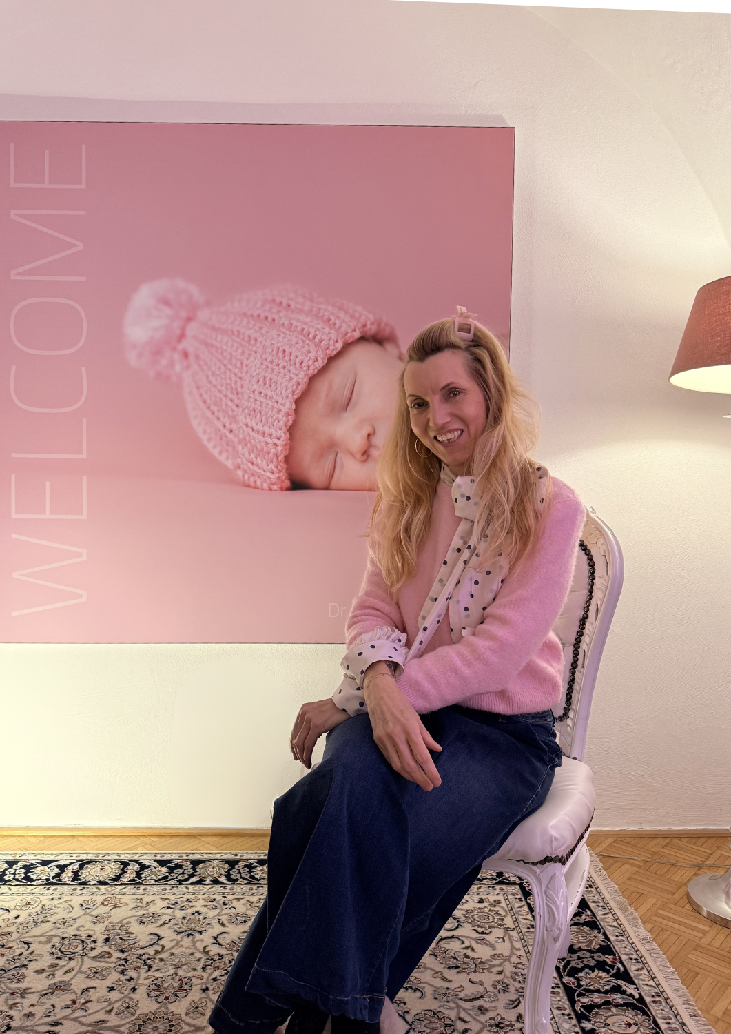 Frau sitzt in einem weißen Stuhl vor einem pinken Wandbild eines schlafenden Babys mit rosa Mütze.