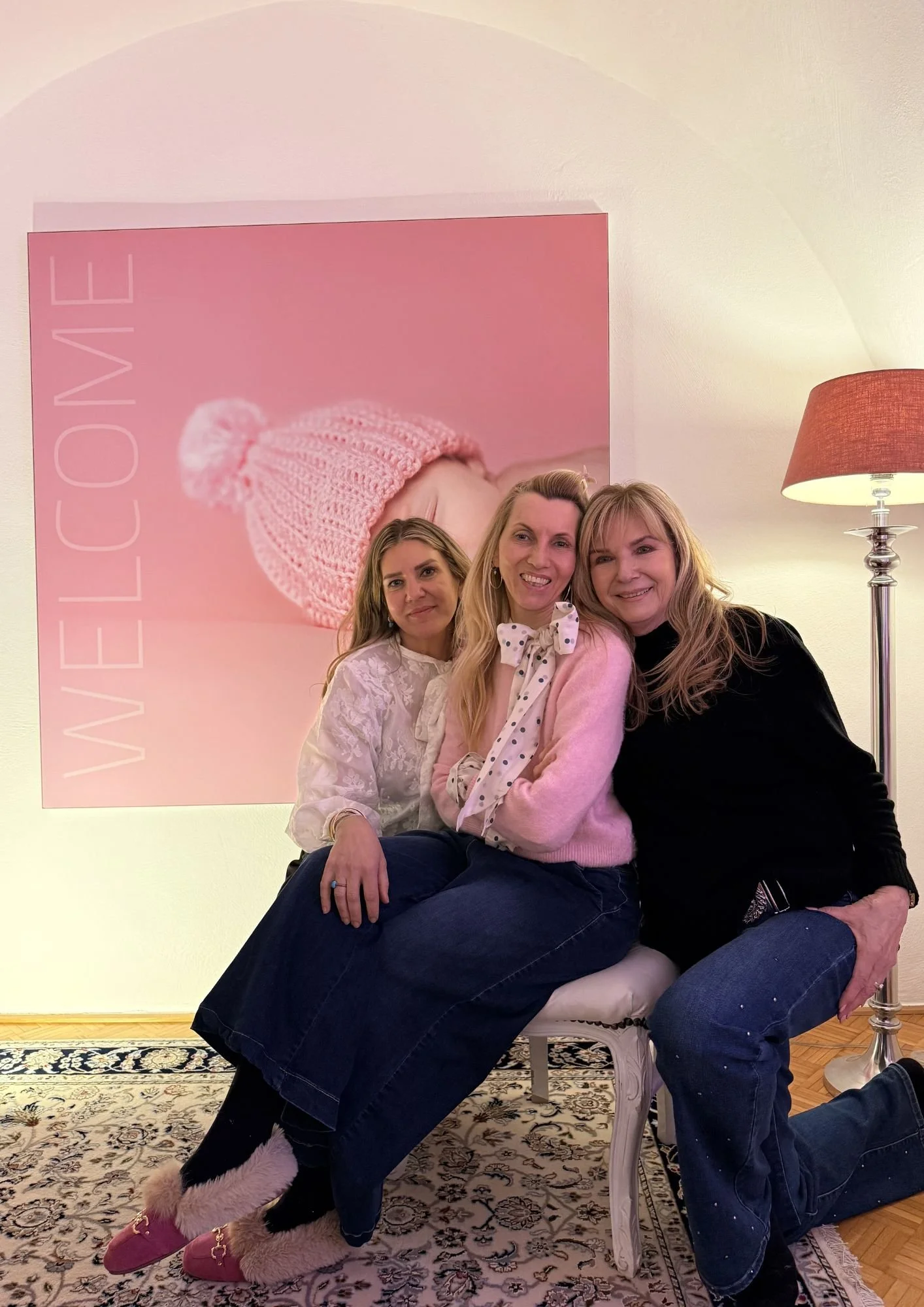 Drei Frauen sitzen eng beieinander auf einem Stuhl vor einem pinken Wandbild mit einem Babybild und der Aufschrift "Willkommen".