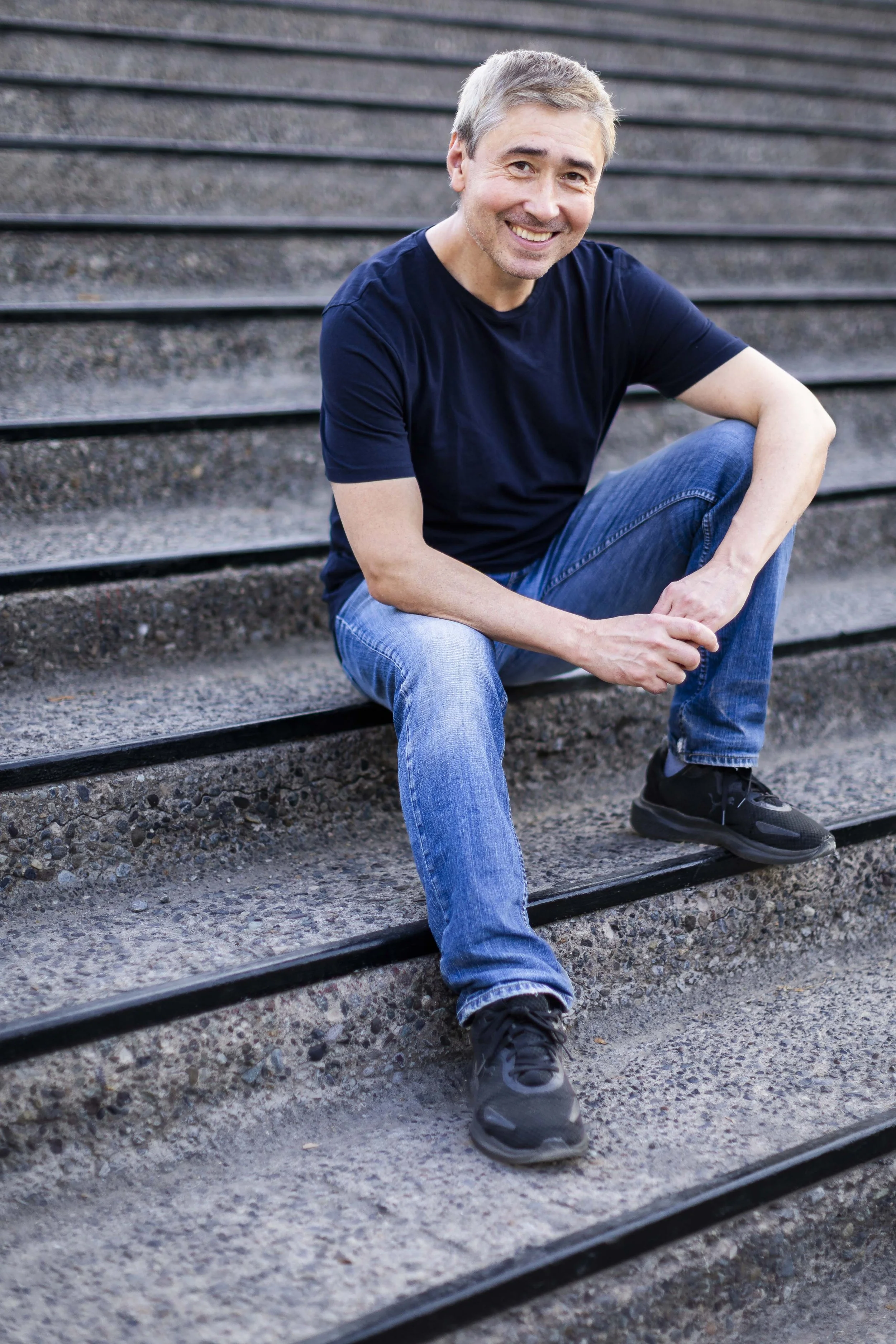 A middle-aged man with gray hair and a beard sitting on outdoor steps, smiling into the camera, wearing a navy blue t-shirt, blue jeans, and black sneakers.