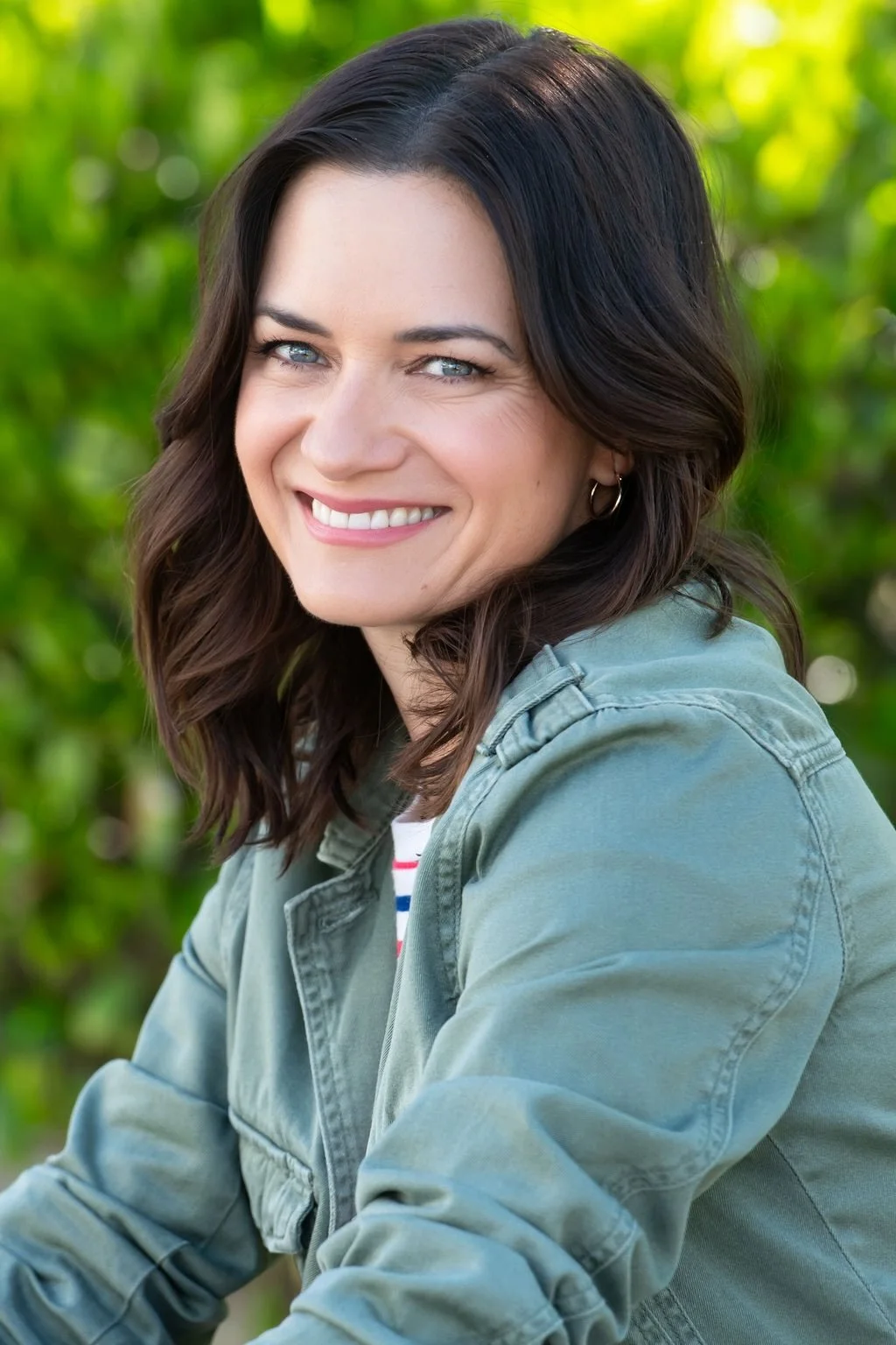 A smiling woman with wavy brown hair, wearing a green jacket, outdoors with green foliage in the background.