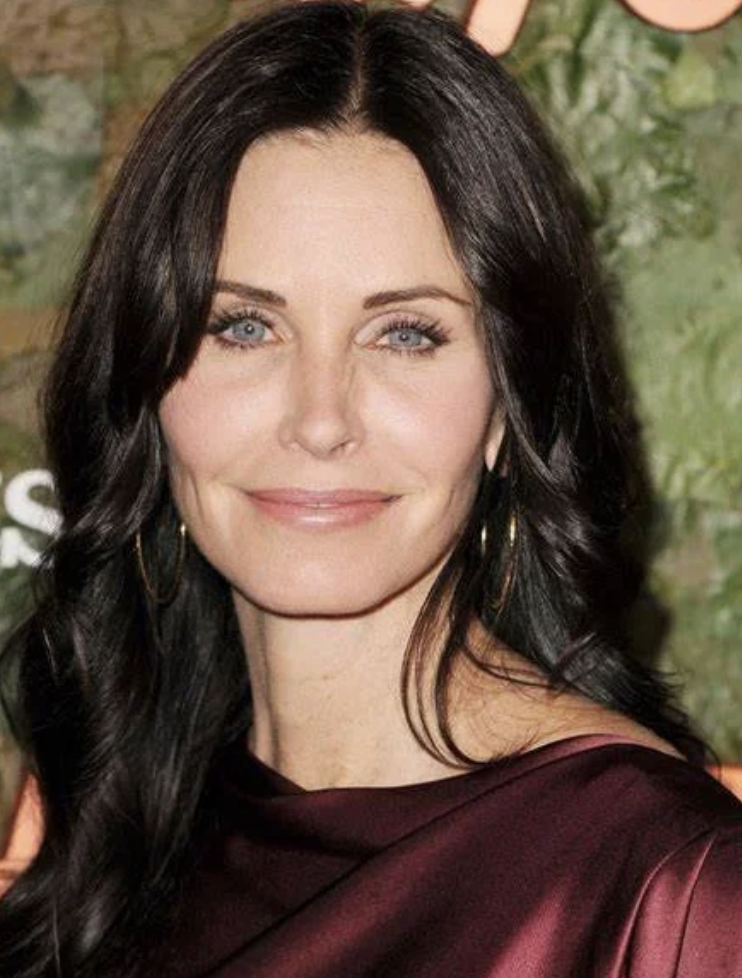Close-up of a woman with long dark hair, blue eyes, and fair skin, smiling gently, wearing hoop earrings and a dark red satin top.