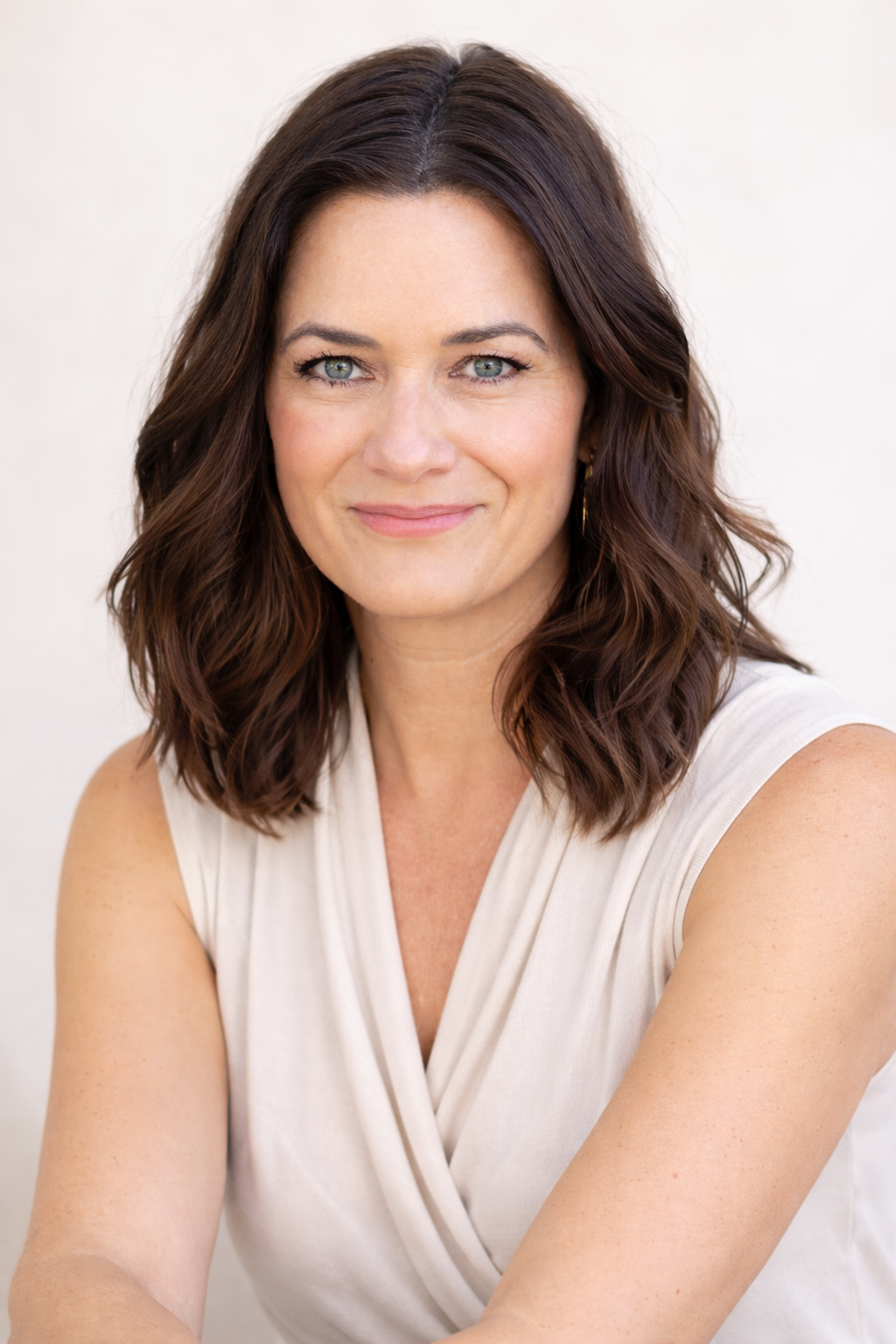 A woman with shoulder-length wavy brown hair, blue eyes, and light skin, wearing a sleeveless beige top, smiling softly, against a plain light background.