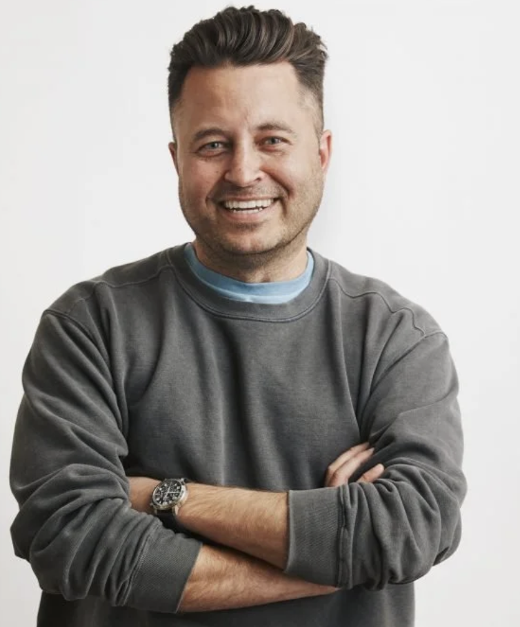 Smiling man with short dark hair, wearing a gray sweatshirt and a wristwatch, standing crossing arms against a plain white background.