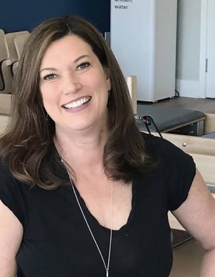 A woman with brown, wavy hair smiling, wearing a black top and a necklace, sitting indoors near a pool table.