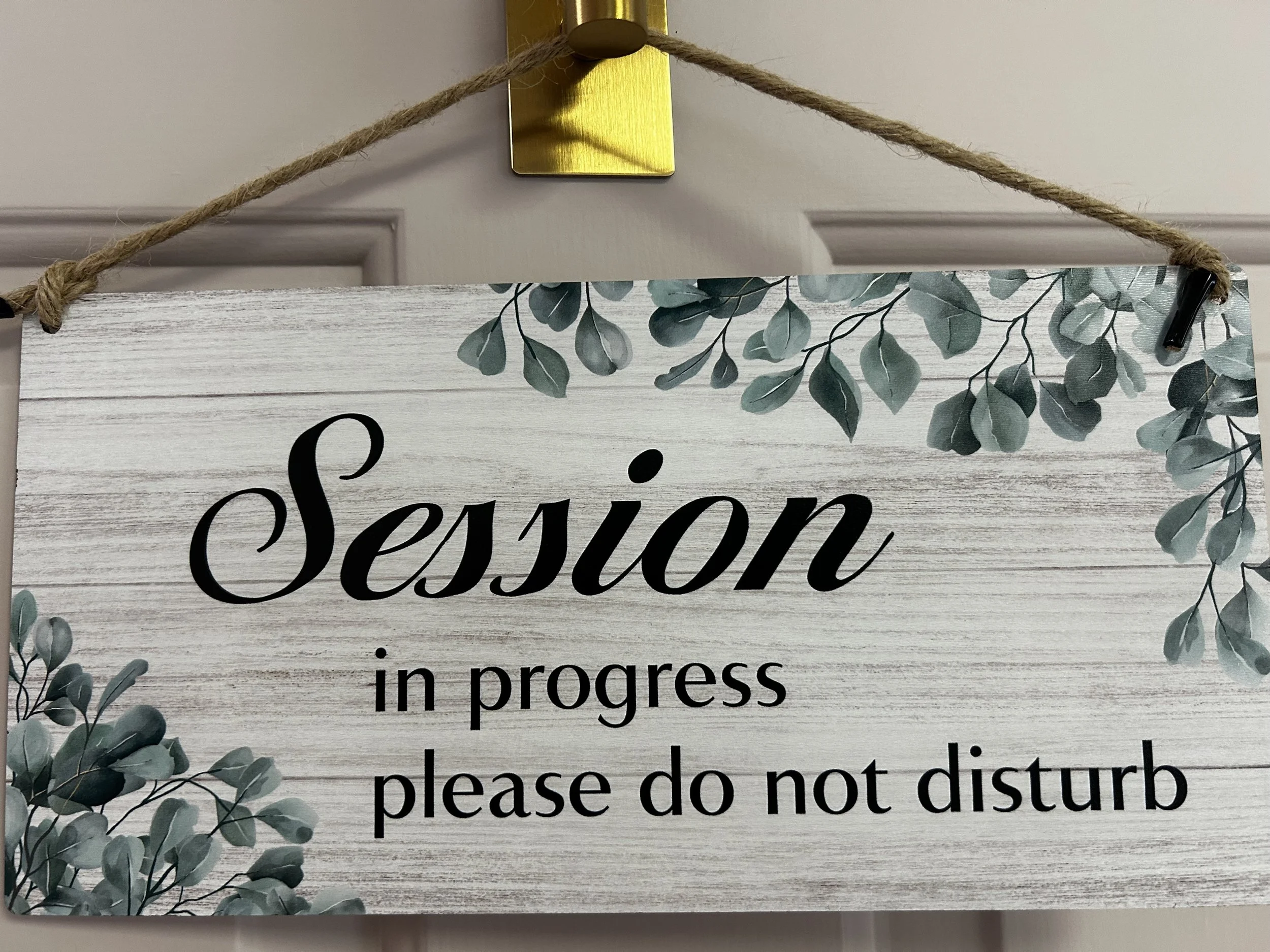 A close-up of a decorative hanging sign with the words "Season in progress" on a white wooden background with greenery, hanging from a rope attached to a brass hook.