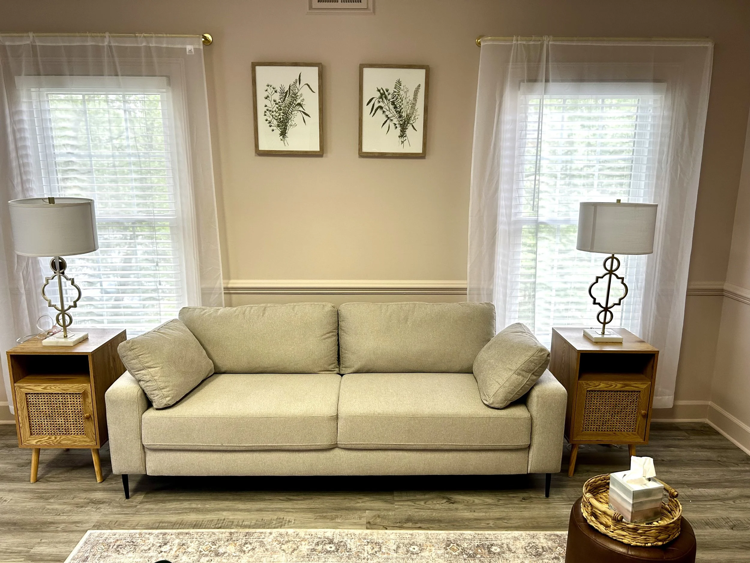Living room with a beige sofa, two wooden side tables with lamps, two large windows with white curtains, wall art of botanical illustrations, and a decorative basket on the floor.