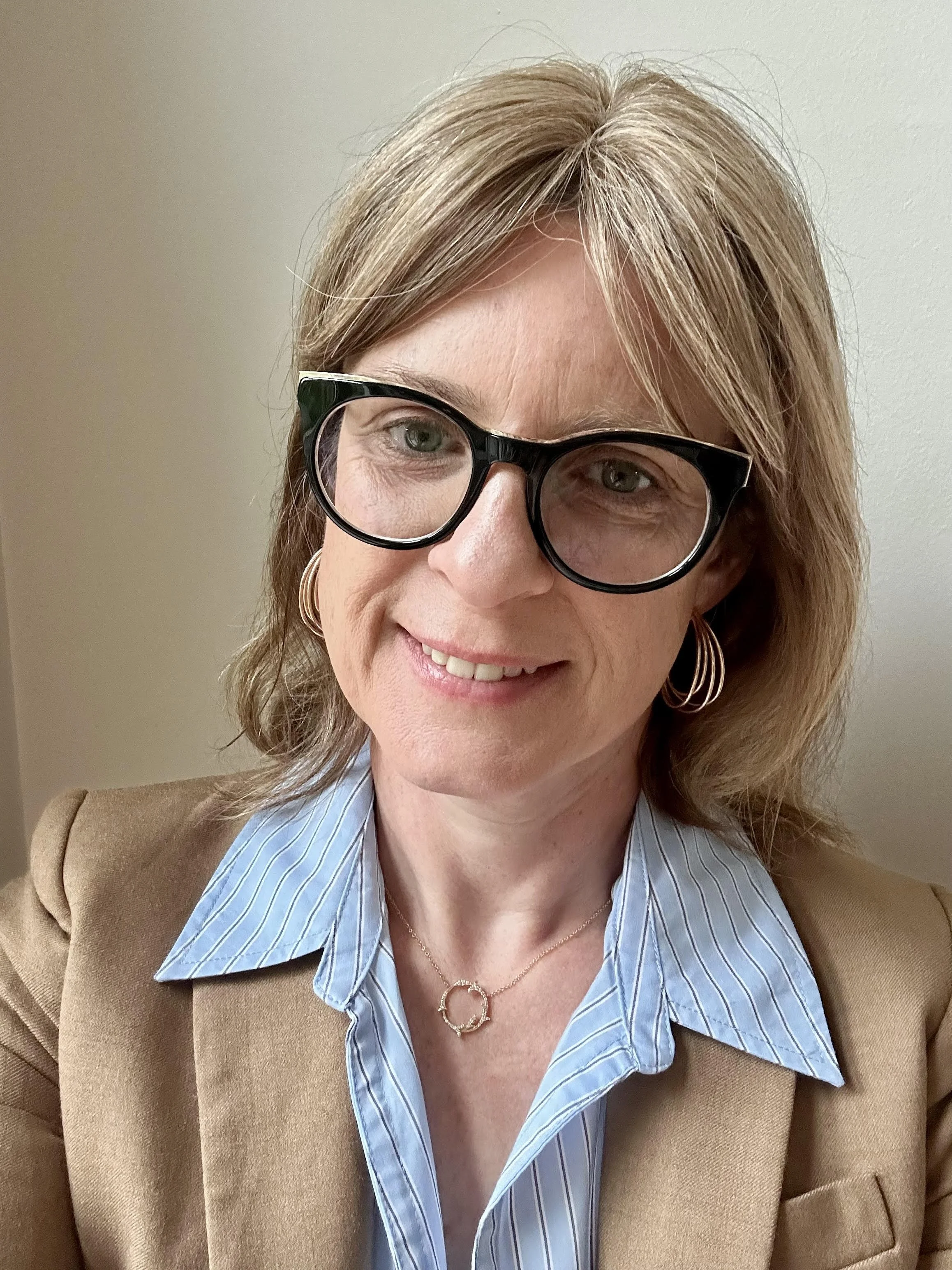 Close-up of a smiling woman with blonde hair, wearing black-framed glasses, gold hoop earrings, a cropped gold necklace, a blue striped shirt, and a tan blazer, standing against a beige wall.