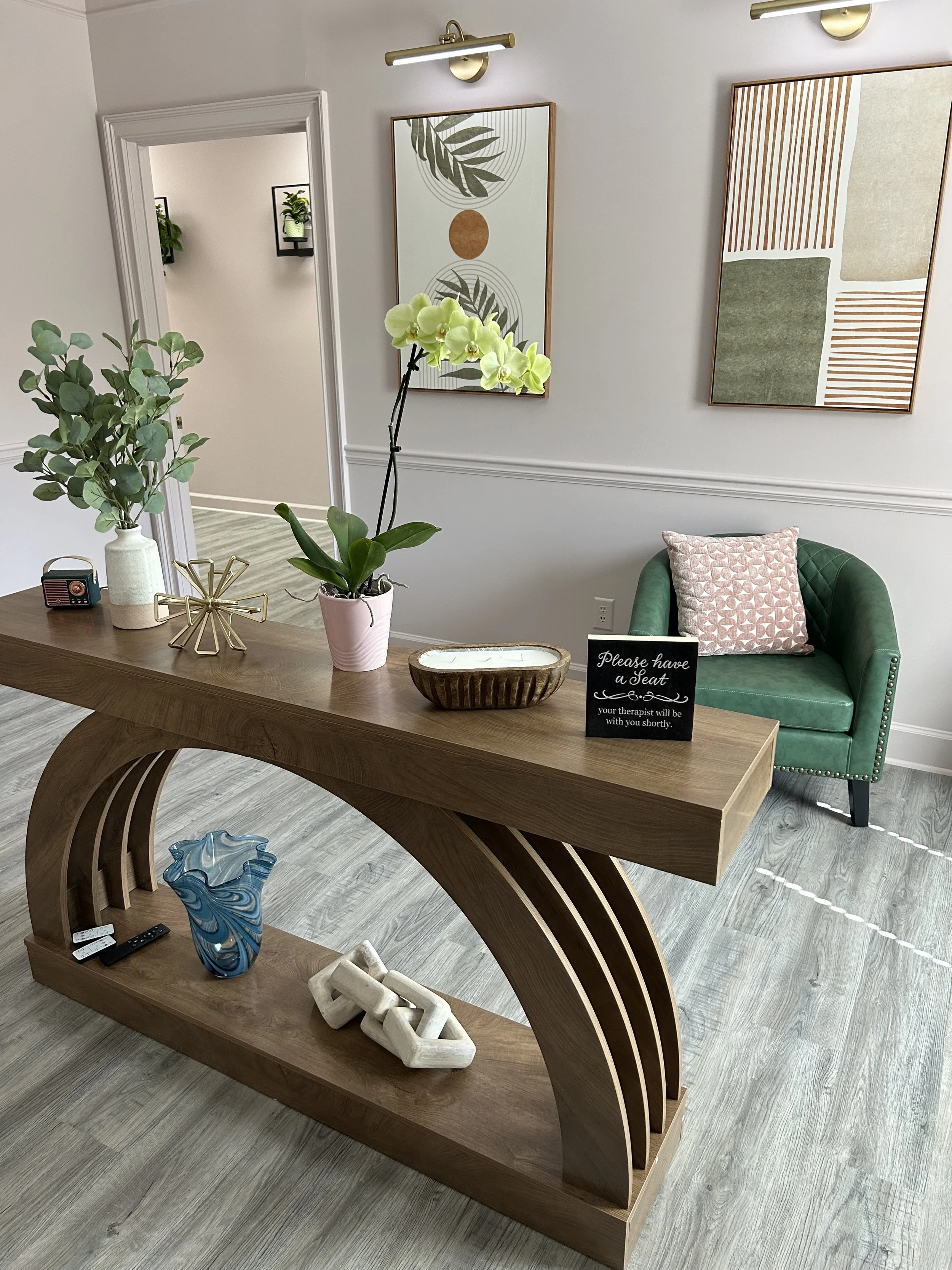 A modern lobby space with a wooden table, decorative objects including a blue and white marbled vase, white geometric sculptures, a pink flowerpot with green orchids, and a small black sign that says, 'Please have a seat, your therapist will be with you shortly.' in front of a green upholstered armchair with a pink patterned pillow. The wall features two abstract botanical art pieces and gold wall sconces. There is a doorway leading to a hallway with wall-mounted plant shelves.
