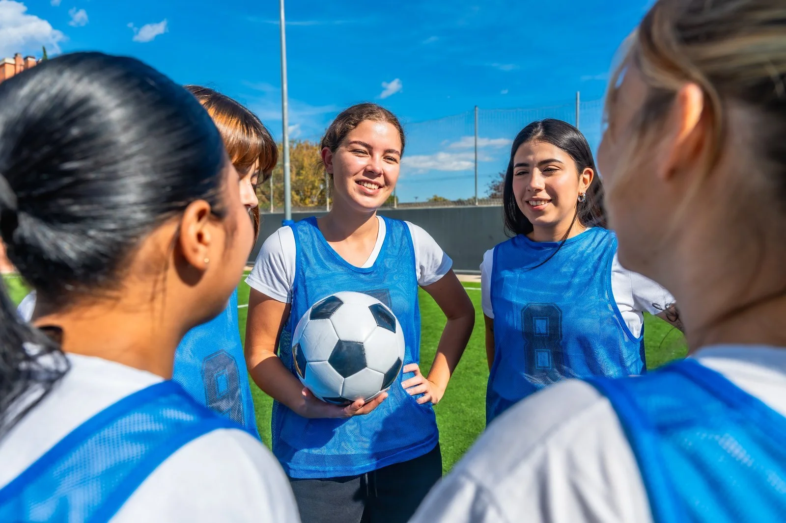 Five girls in blue training vests gathered on a soccer field, with one holding a ball.