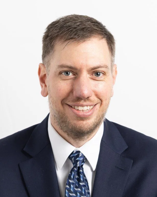 Professional headshot of a man in a dark suit, white shirt, and patterned tie, smiling with short brown hair against a plain white background.