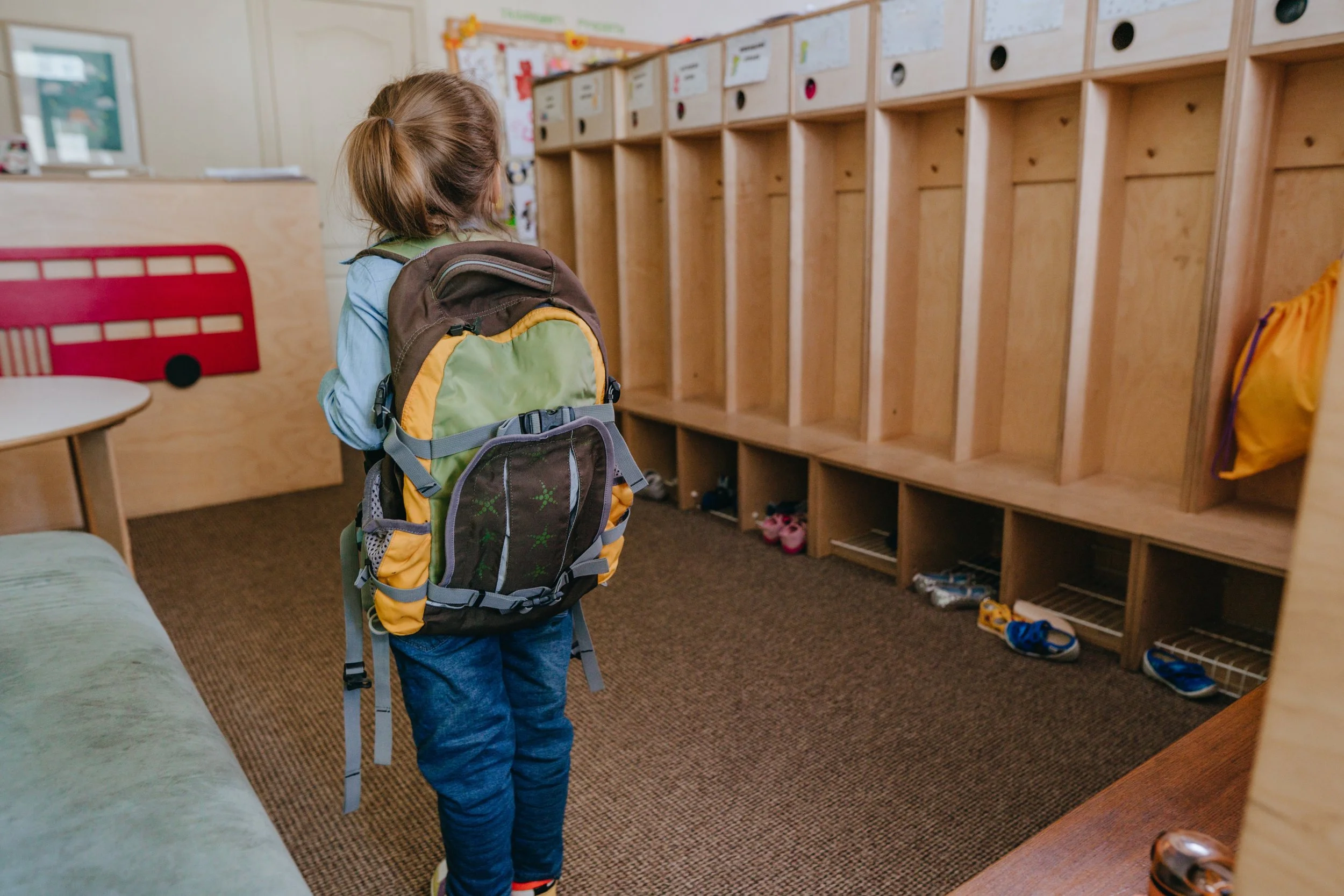A child wearing a backpack stands inside a classroom beside a row of empty cubbies.