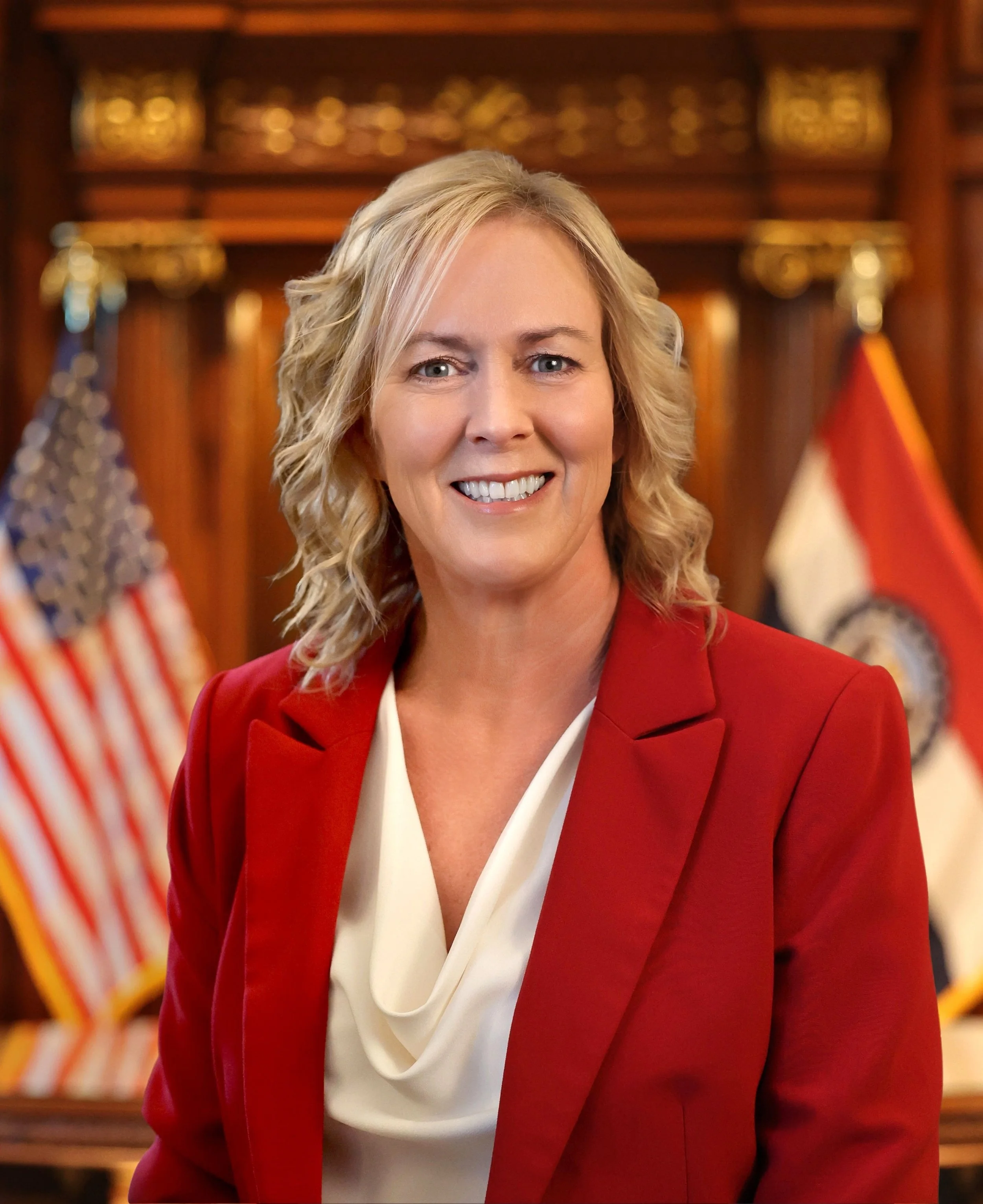 Missouri First Lady Claudia Kehoe. A smiling blonde woman in a red blazer and white blouse standing in a formal setting with American and state flags in the background.
