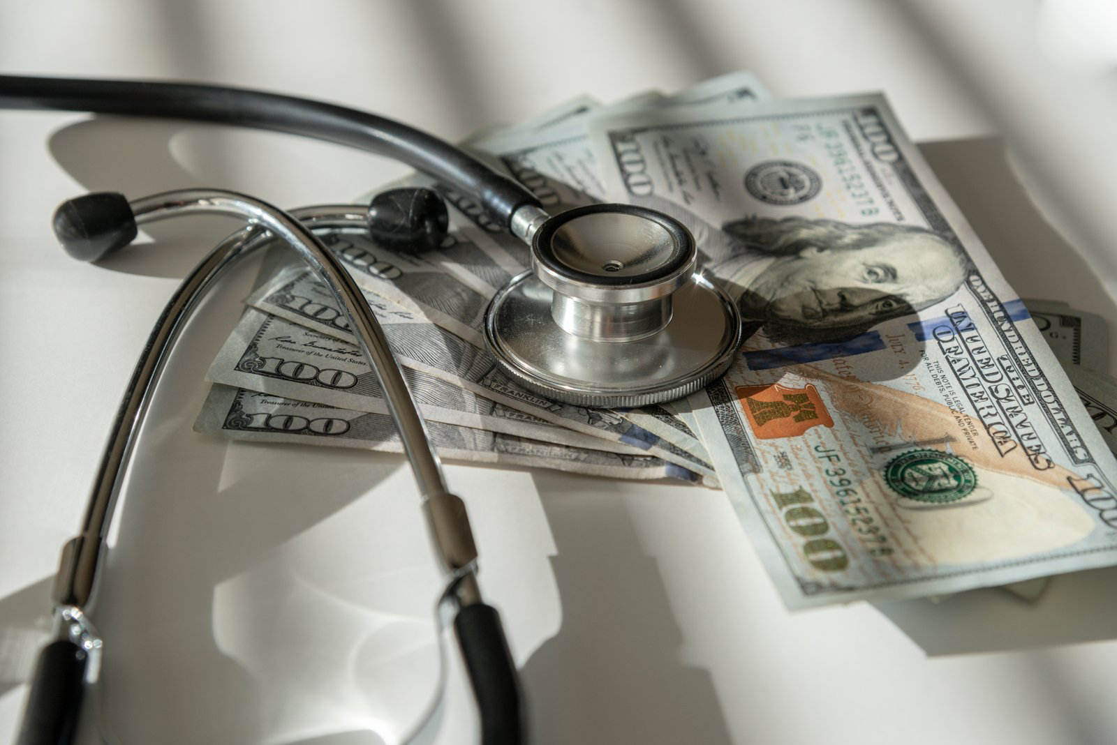 A black stethoscope resting on U.S. one-hundred-dollar bills on a white surface.