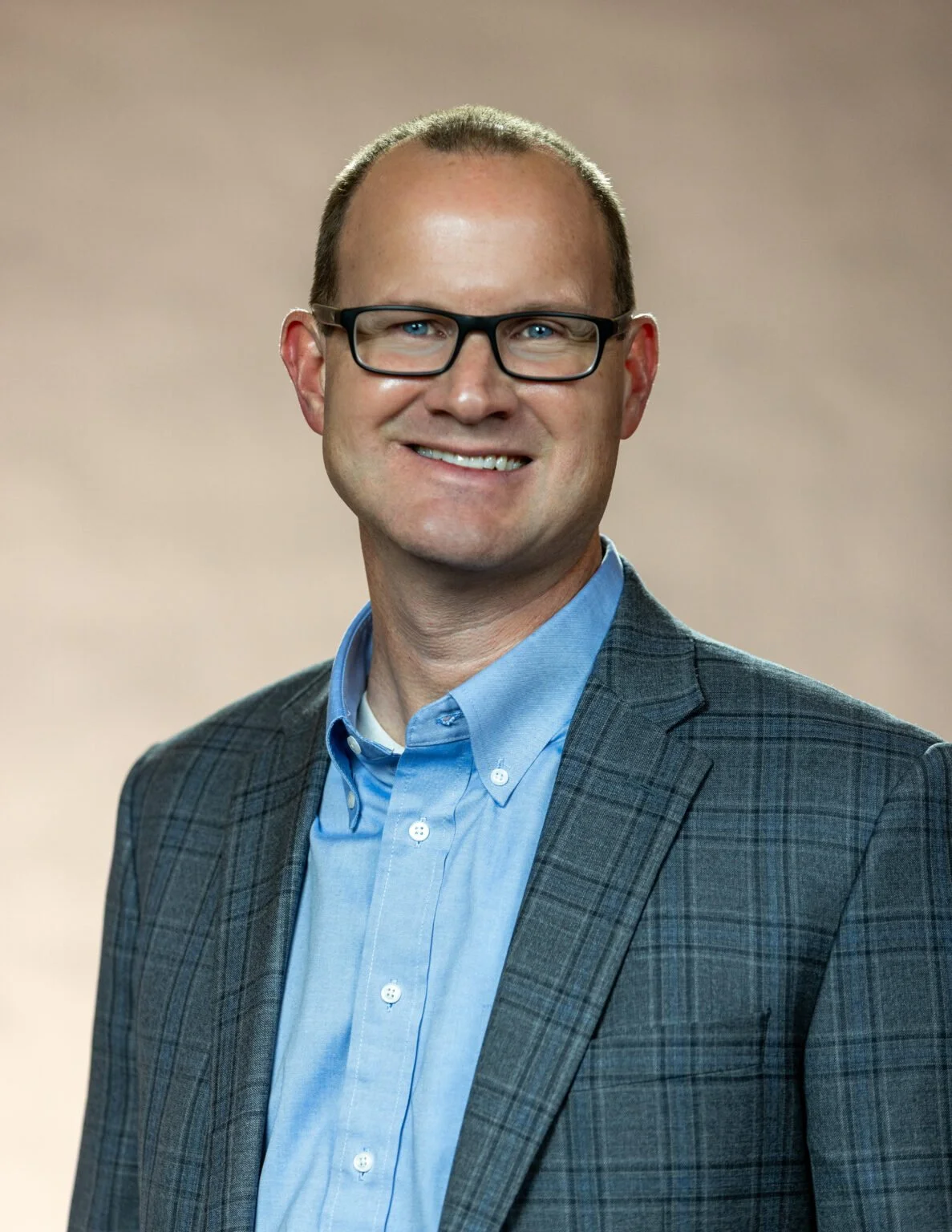 A man with glasses, wearing a blue shirt and a gray plaid blazer, smiling against a neutral background.