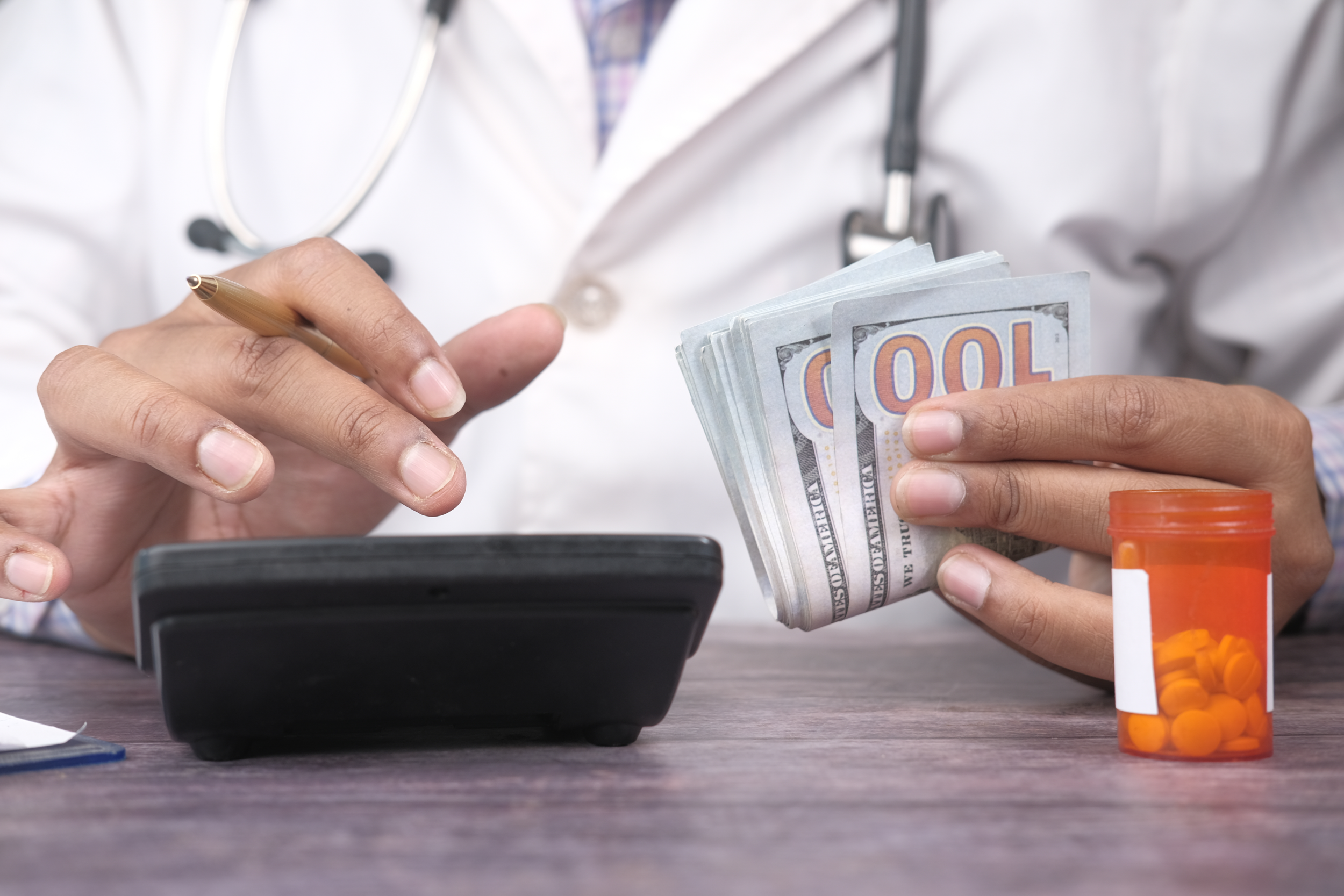 Hands in a white lab coat counting cash beside a calculator and a bottle of pills