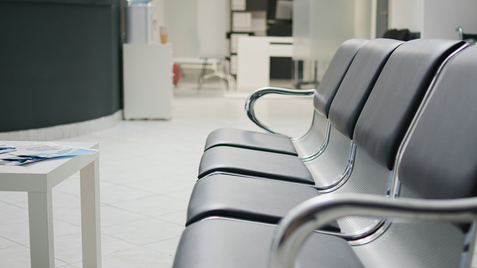 Empty black chairs with metal armrests beside a table of leaflets in a medical waiting room.