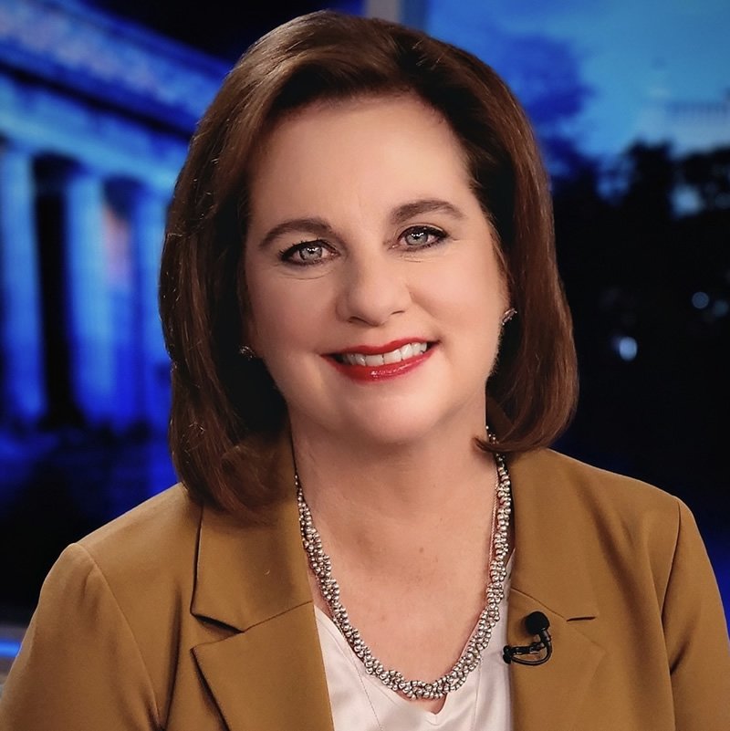 A woman with shoulder-length brown hair, wearing a tan blazer and jewelry, smiling in front of a nighttime background featuring illuminated columns.
