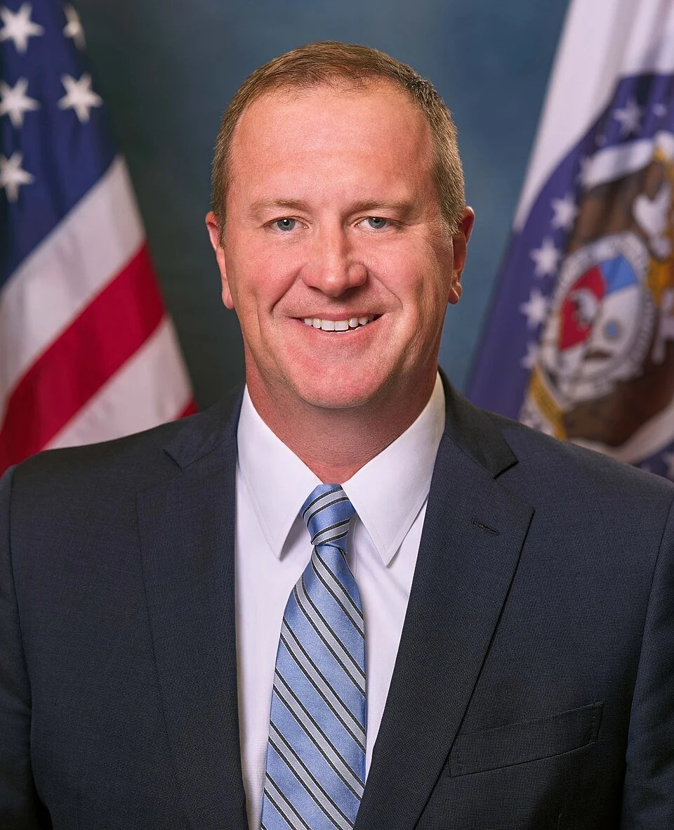 U.S. Senator Eric Schmitt. A smiling man in a suit and tie is standing in front of an American flag and a state flag, with a blue background.