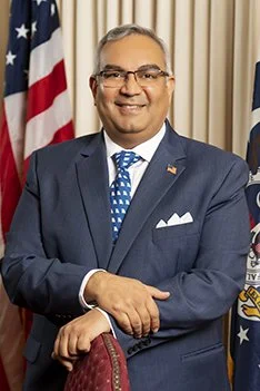Missouri State Treasurer Vivek Malek. A smiling man in a blue suit and blue patterned tie standing in front of American flags and a beige curtain.