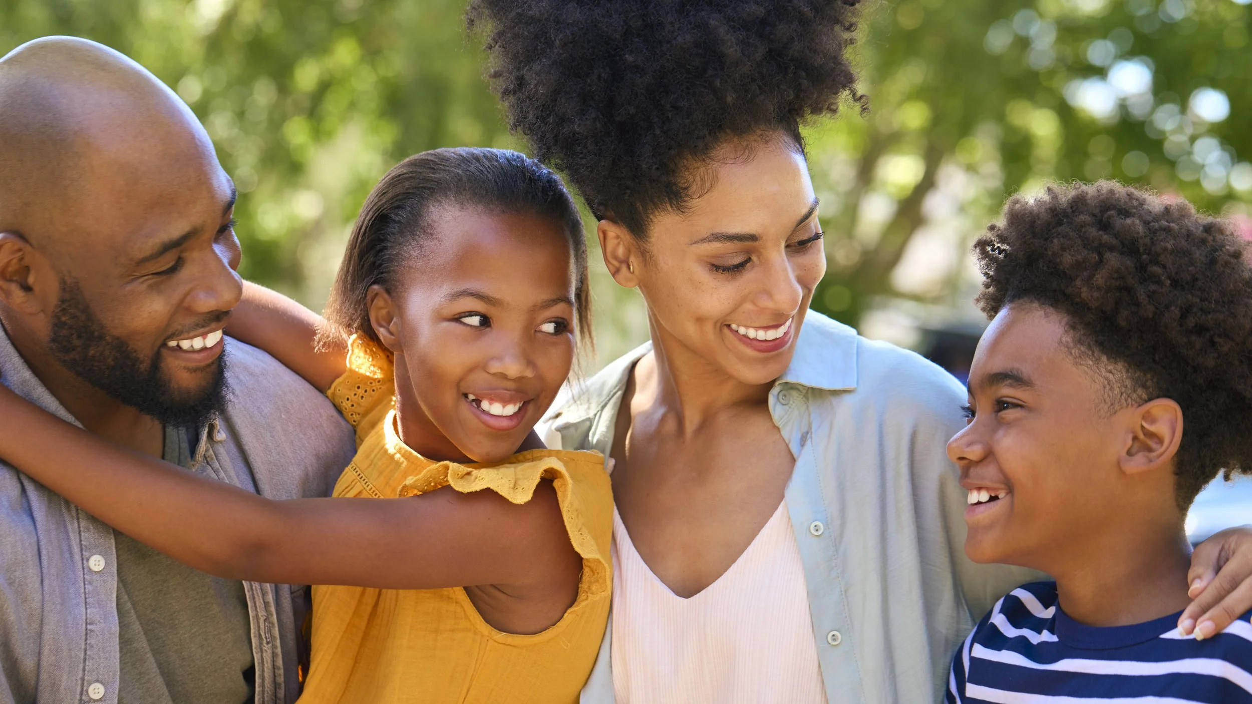 A happy family of four, including a father, mother, daughter, and son, smiling and enjoying time together outdoors in a park.