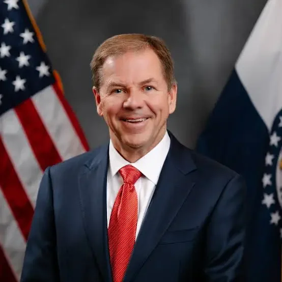 Missouri Lt. Governor David Wasinger. A smiling man in a navy blue suit with a red tie, standing in front of an American flag and a navy blue flag with white stripes.