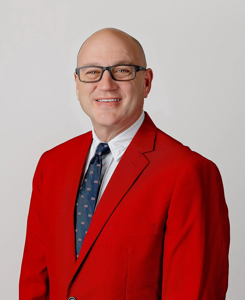 Missouri Secretary of State Denny Hoskins. A man wearing a red blazer, white shirt, and navy tie with small American flags, smiling and wearing glasses, standing against a plain white background.