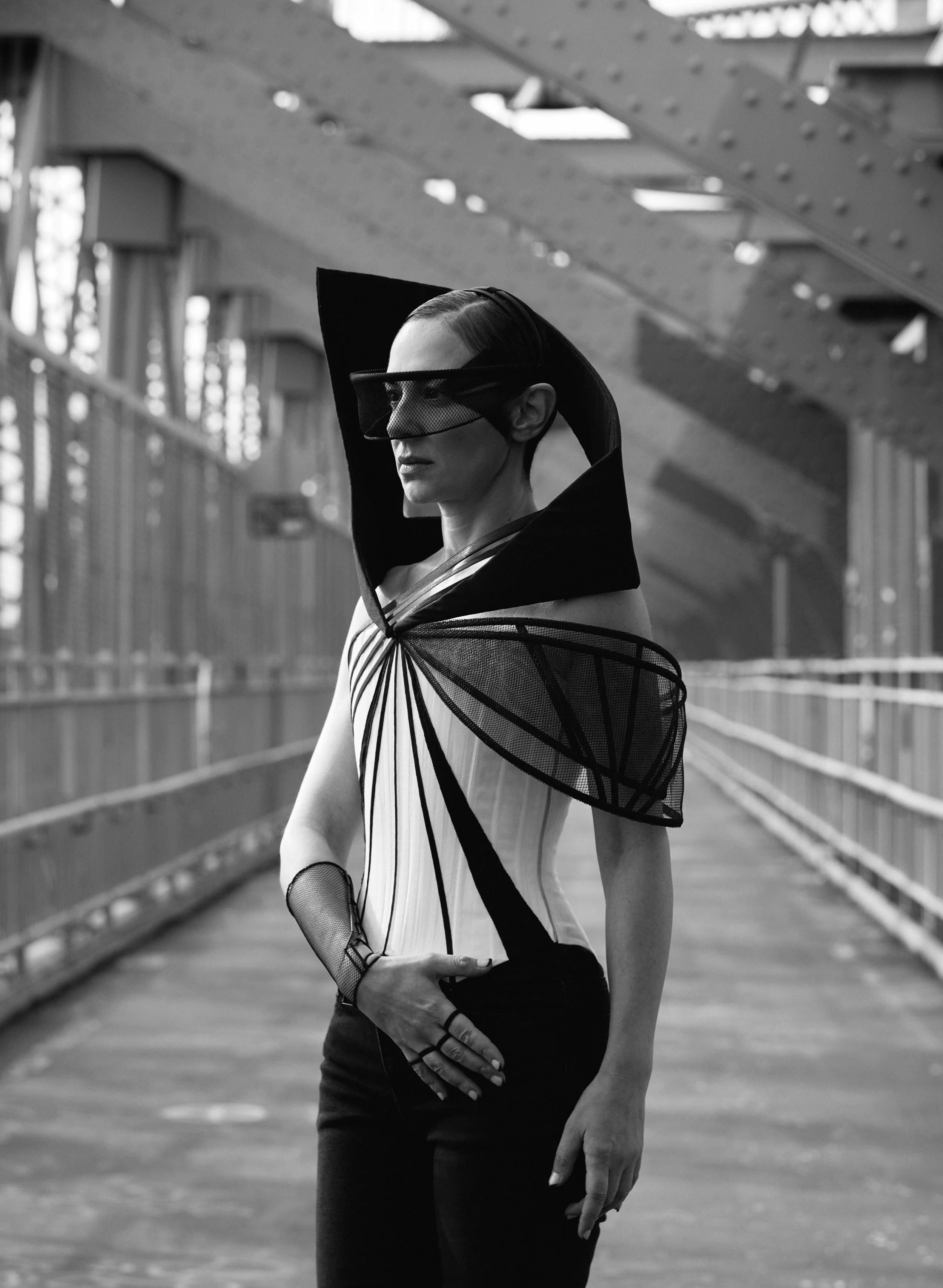 Nicky Conti in the Ironick look framed by the steel beams of the Williamsburg Bridge, wearing a structured corset with diagonal lines and mesh detailing around one arm