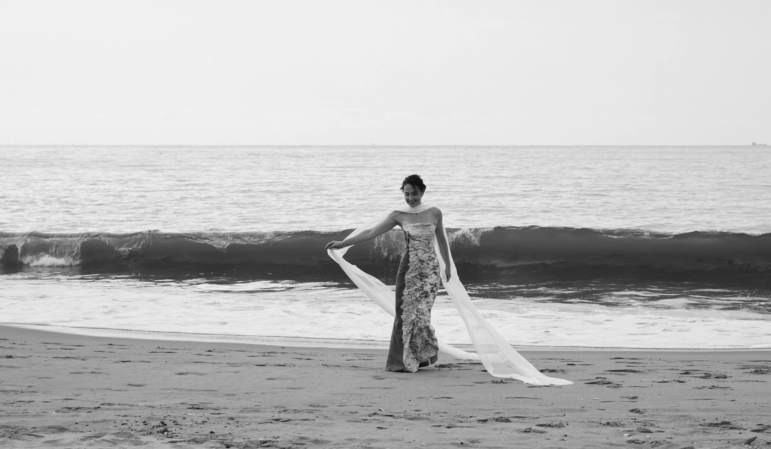 Nicky Conti in the distance on Coney Island Beach, wearing the Memoria couture piece and moving a long scarf as a wave rises behind her