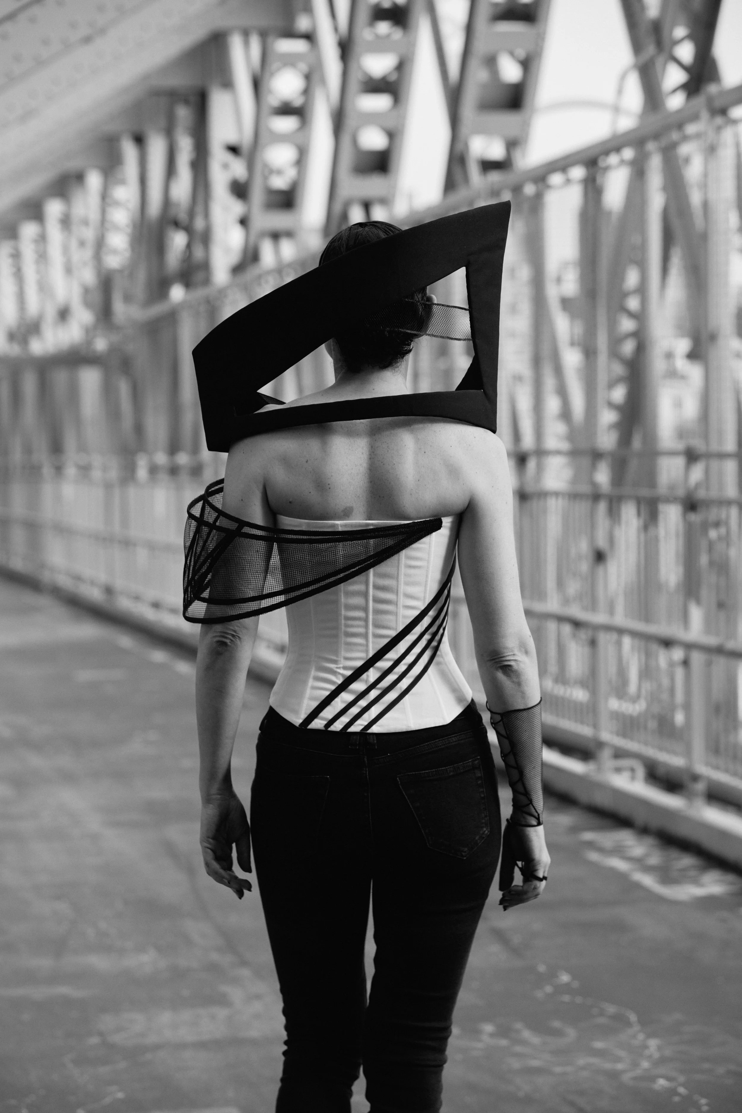 Back view of Nicky Conti walking away on the Williamsburg Bridge wearing the Ironick look, showing the sculptural headpiece and fitted corset structure