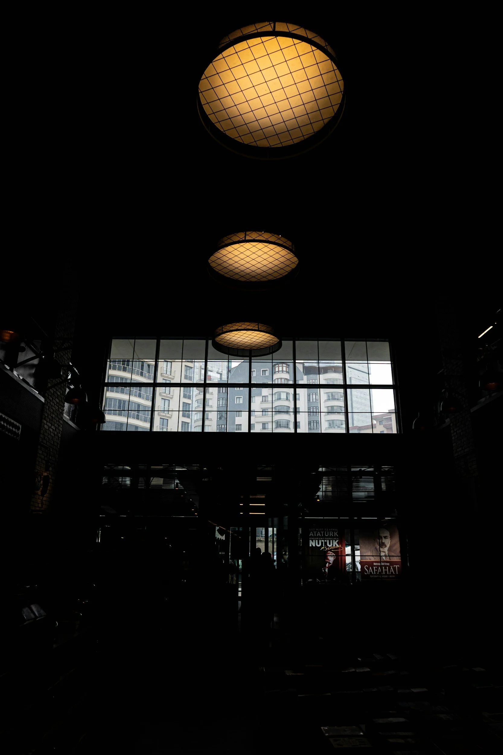 Interior of a building with large windows, ceiling lights, and high-rise buildings visible outside.