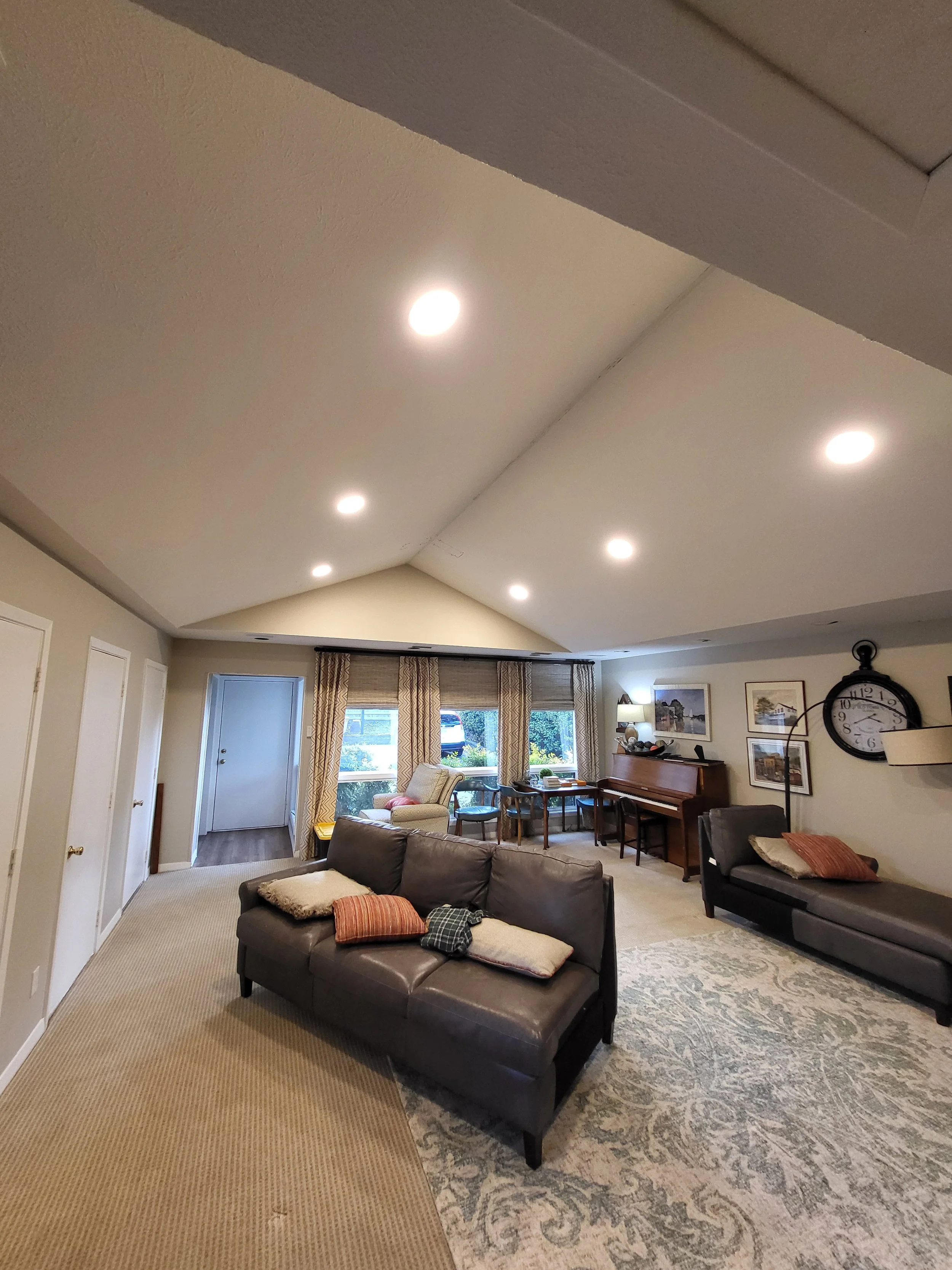 Living room with high vaulted ceiling, multiple recessed lights, beige walls, a gray sectional sofa with pillows, a patterned area rug, a piano, artwork on the walls, large window with curtains, and a door leading outside.