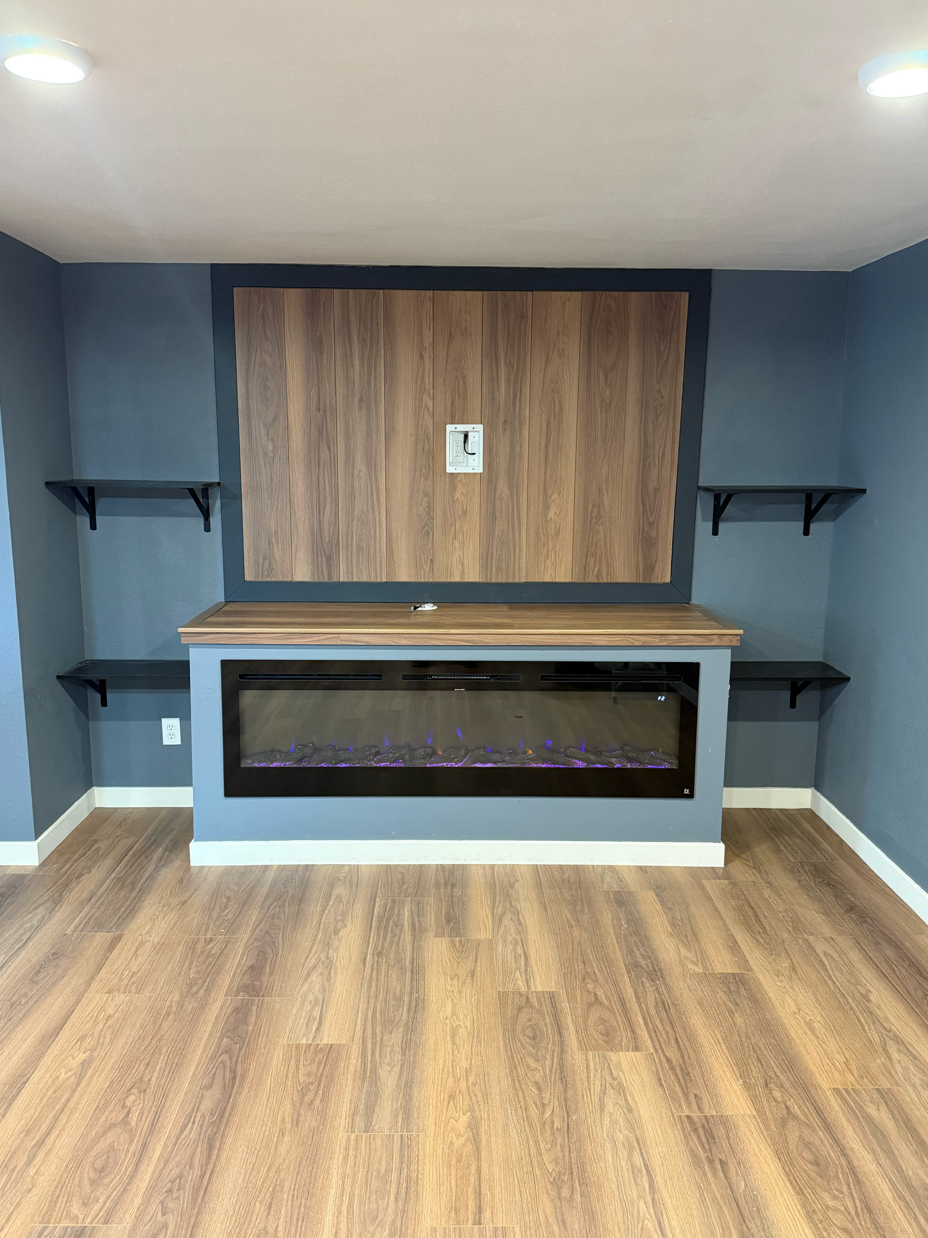 Empty room with wood floor, painted blue wall, and three black wall-mounted shelves. In the center, there is a wooden table with a built-in electric fireplace with purple flames. A beige ceiling with recessed lighting.