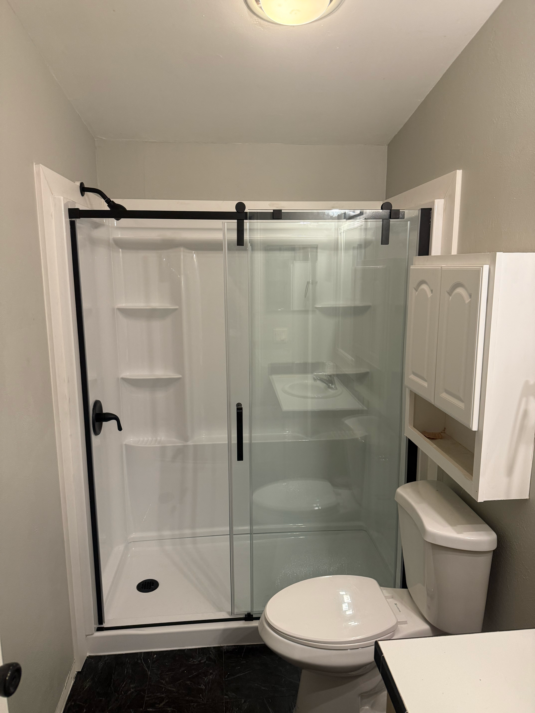 A white bathroom with a glass shower enclosure, a white toilet, and white cabinets above the sink. The shower has built-in shelves and a sliding glass door with black accents.