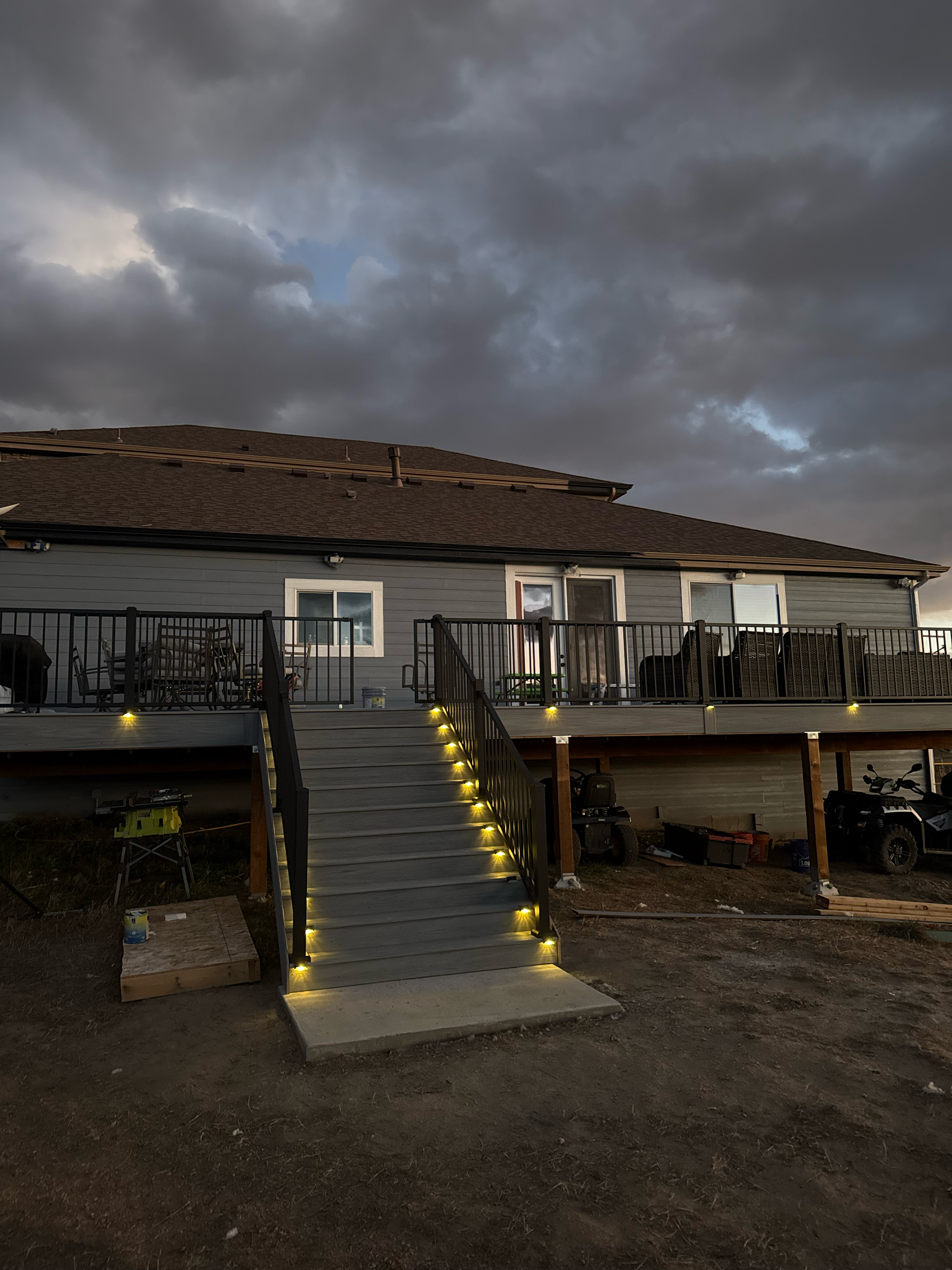 A newly built house with a deck and stairs, illuminated by yellow lights, under a dark, cloudy sky.
