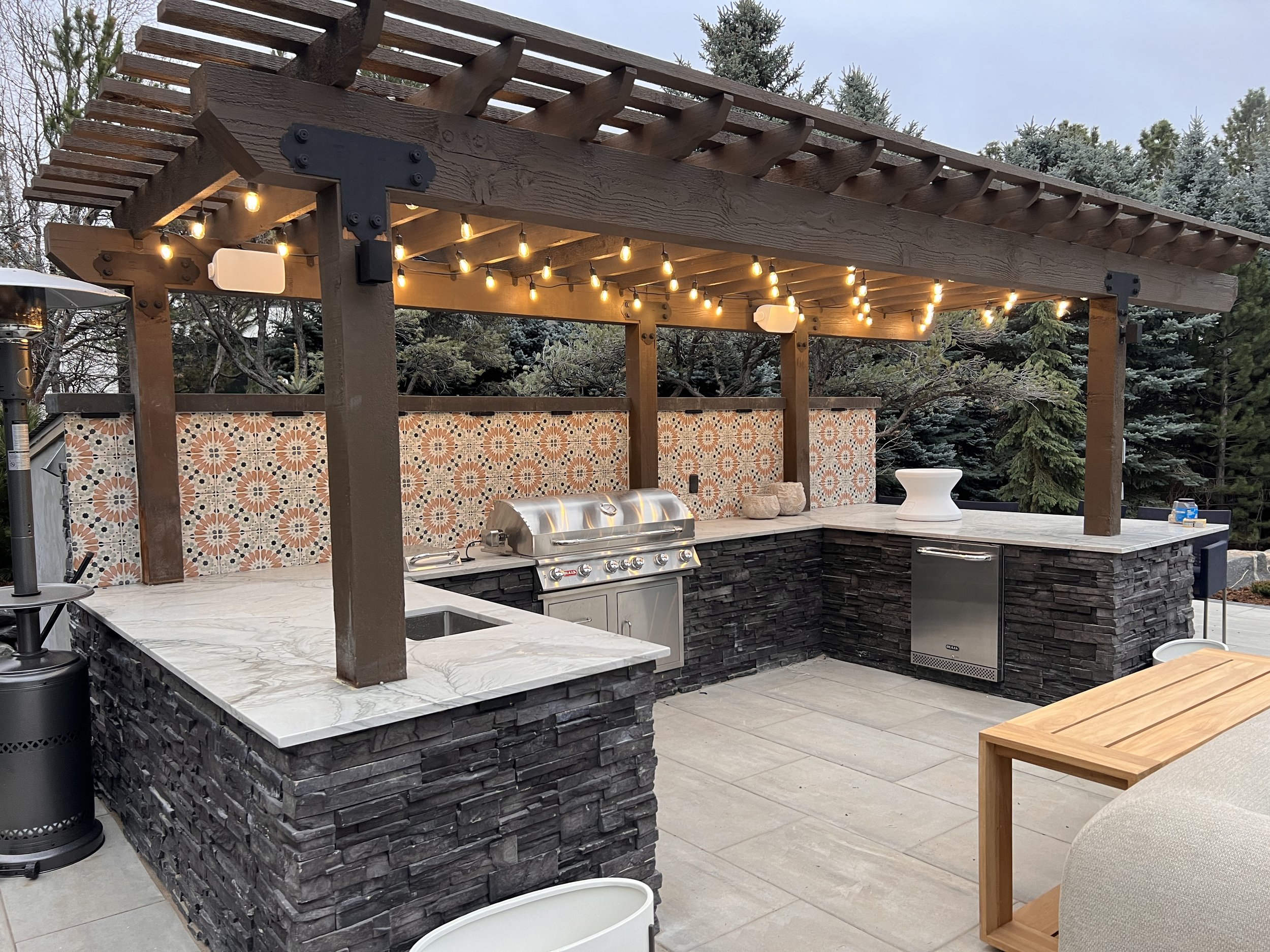 Outdoor kitchen with a bar area, string lights, a built-in grill, a marble countertop, and a decorative tile backsplash, surrounded by trees.