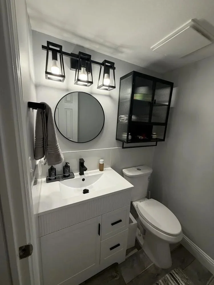Modern bathroom with a white vanity, round mirror, black fixtures, wall-mounted cabinet with glass doors, decorative light fixture, and a toilet.
