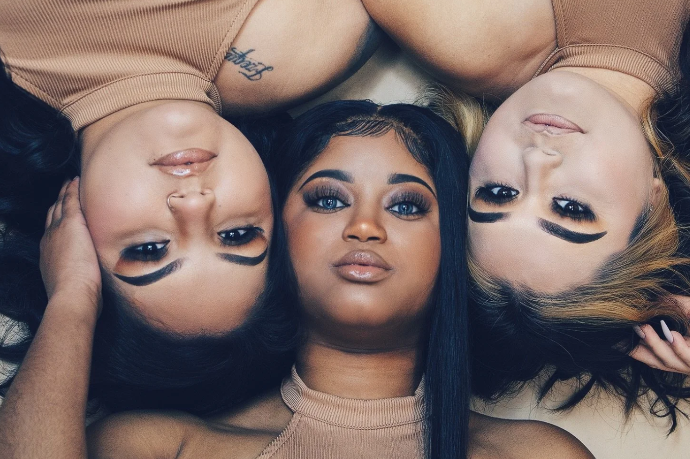 Three women lying on a surface with their heads together, looking up at the camera. They have makeup on with bold eyebrows and lashes, and their hair is styled differently.