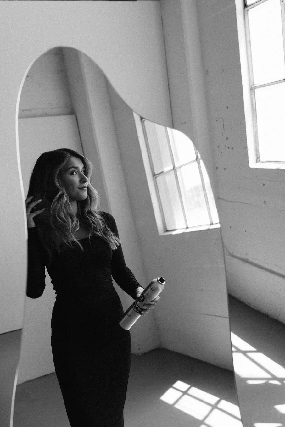 A woman with wavy hair in a black dress standing in front of a large irregularly shaped mirror in a room with tall windows, holding a can and looking at her reflection.