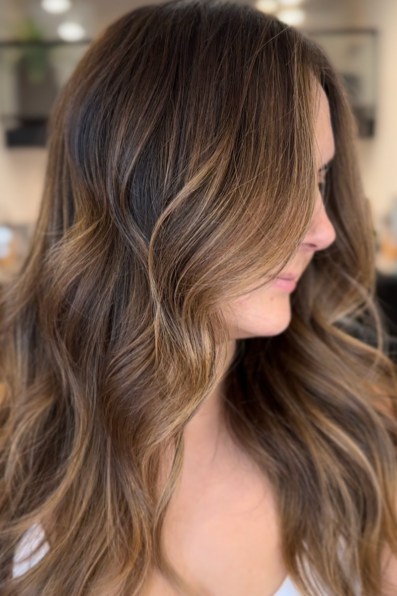 Close-up of a woman's salon hair with caramel highlights and soft waves, seen from the side.