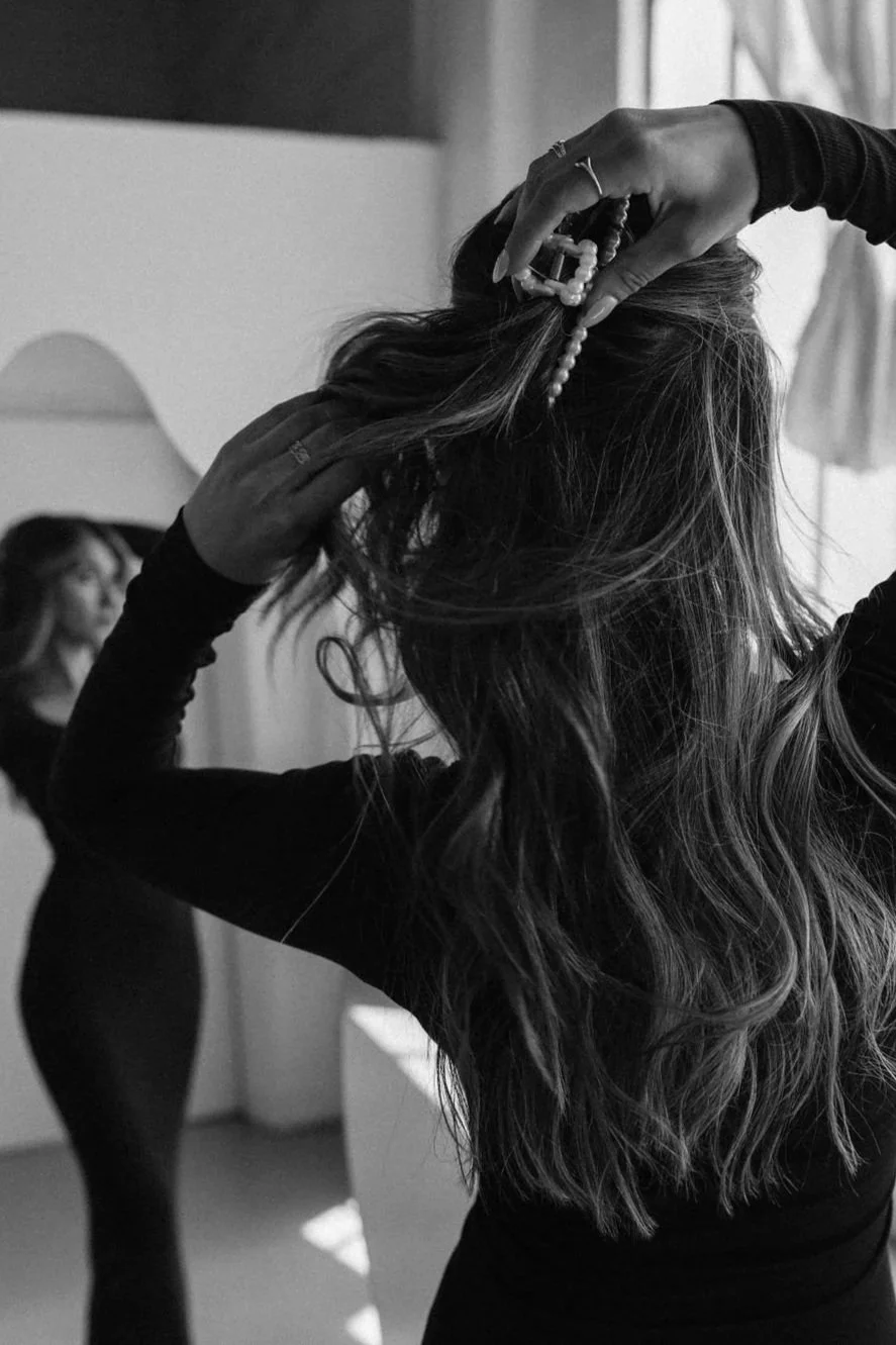 A person with wavy hair is fixing their hair with a hair clip while looking in a mirror, with another woman visible in the background.